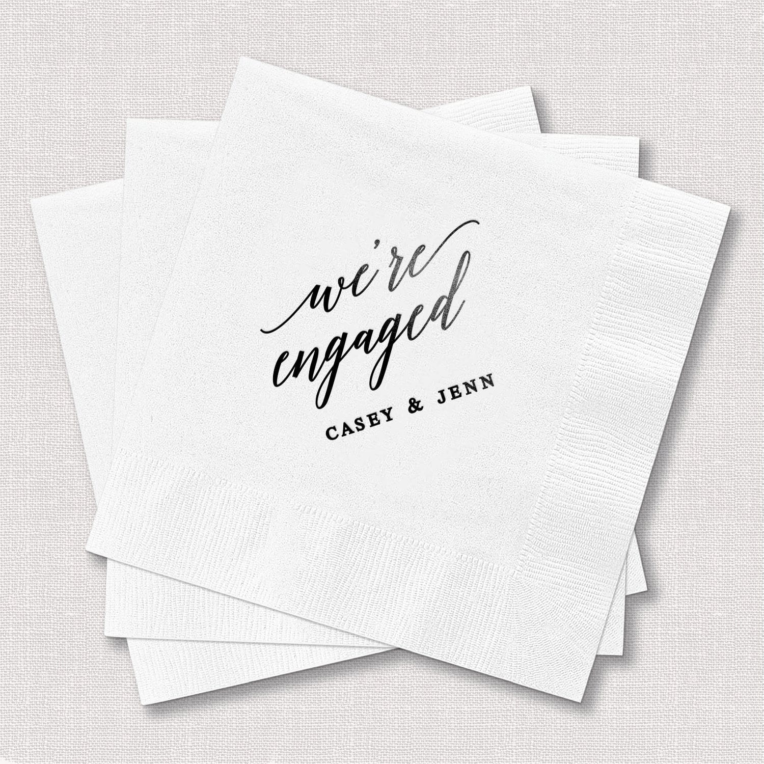 Stack of white napkins with "we're engaged" in script and "CASEY & JENN" personalized in black foil stamping