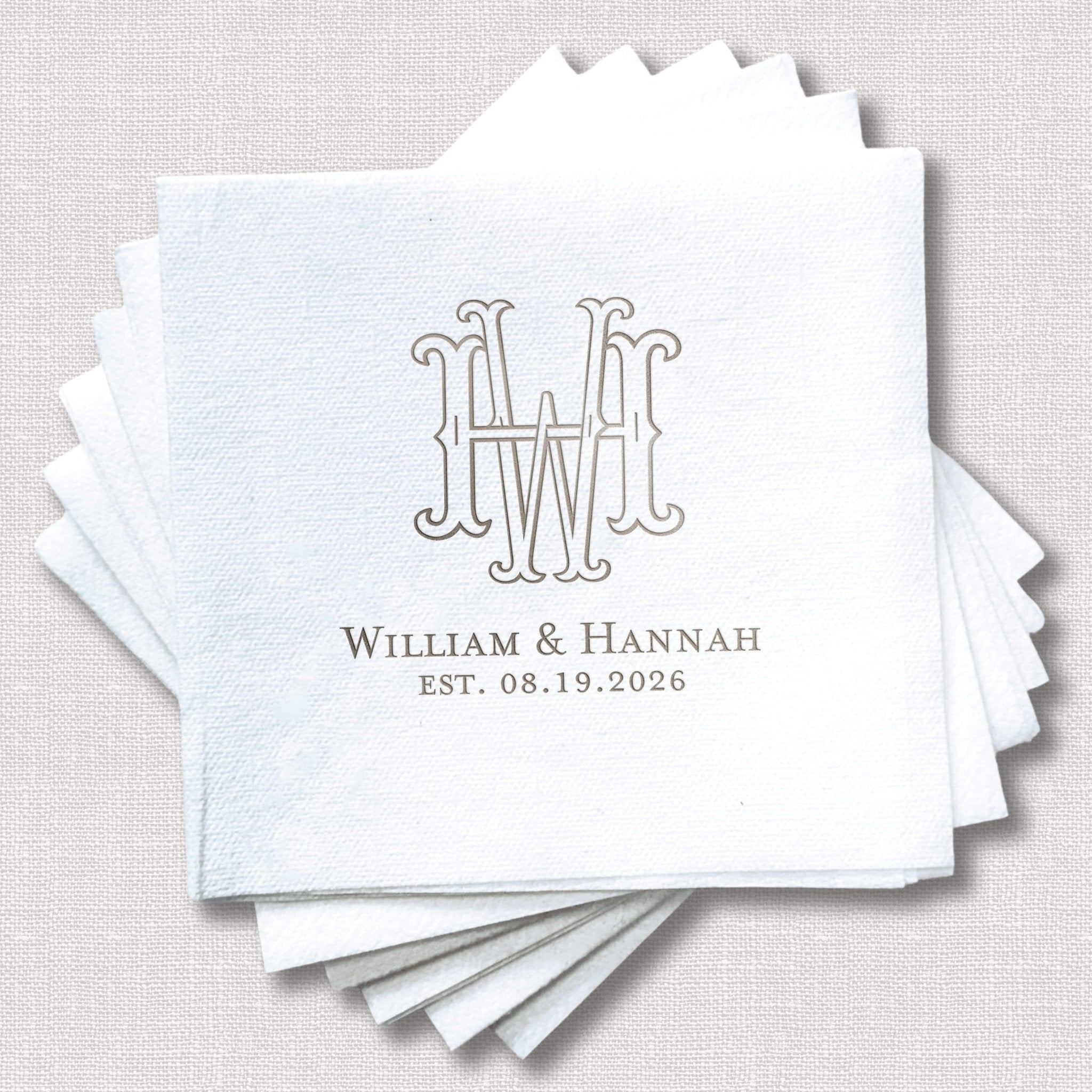 Stack of white linen napkins with interlocking monogram and couple names William & Hannah with wedding date EST. 08.19.2026 in silver foil stamping.
