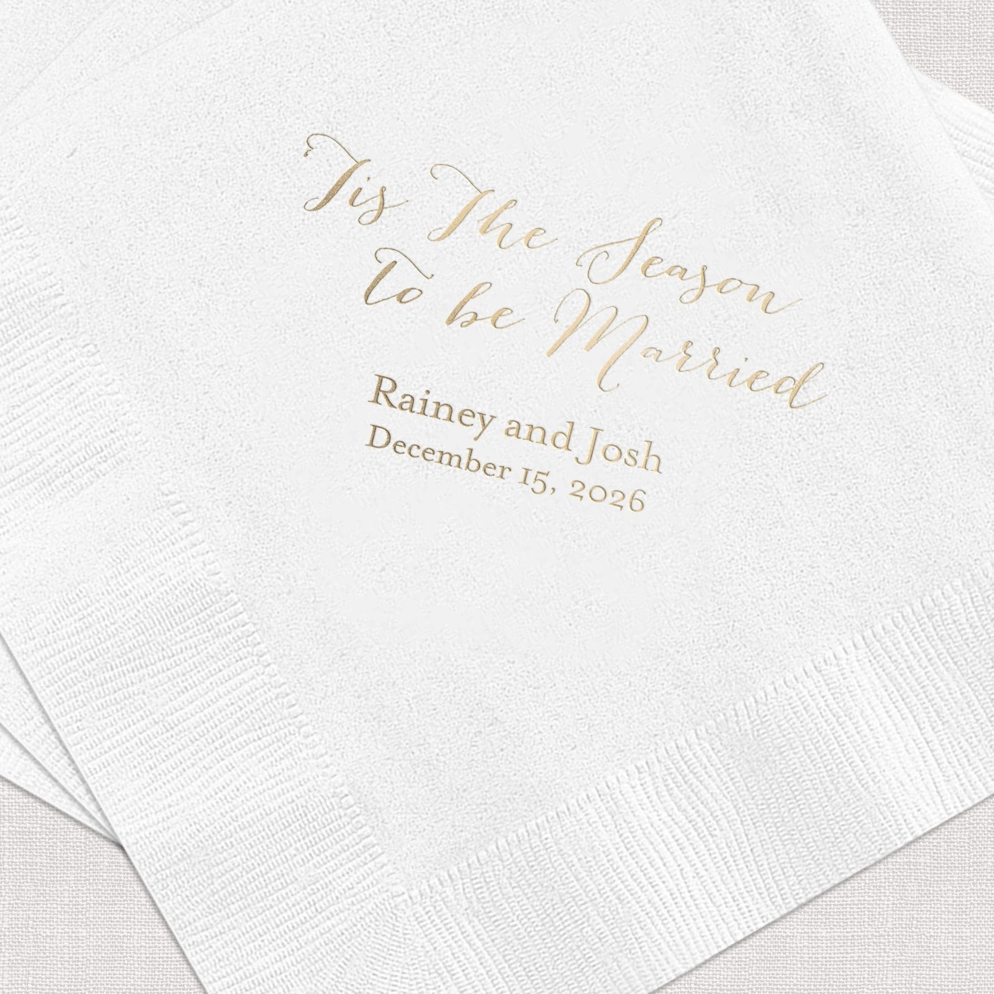 White beverage napkins with gold foil text reading "Tis The Season to be Married" and personalized names "Rainey and Josh" with wedding date December 15, 2026.