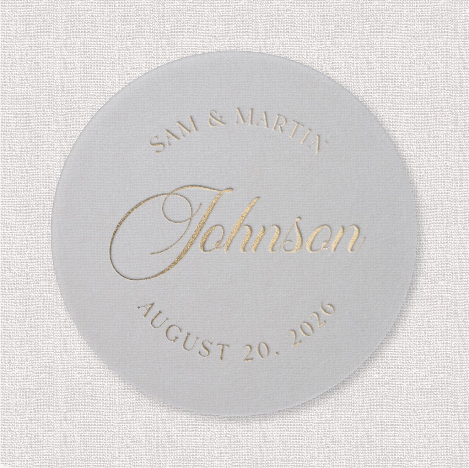 White round coaster with gold foil text reading "SAM & MARTIN" at top, "Johnson" in script in center, and "AUGUST 20, 2020" at bottom.