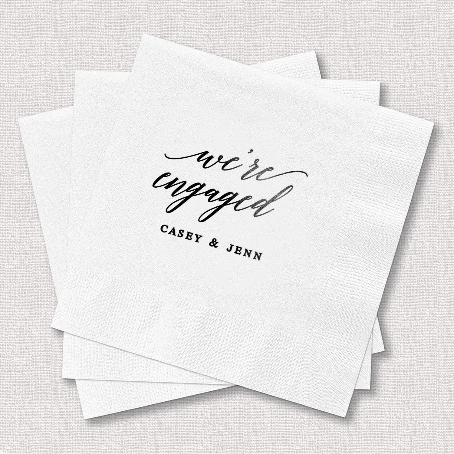 White cocktail napkins with "we're engaged" script and "CASEY & JENN" printed in black foil stamping
