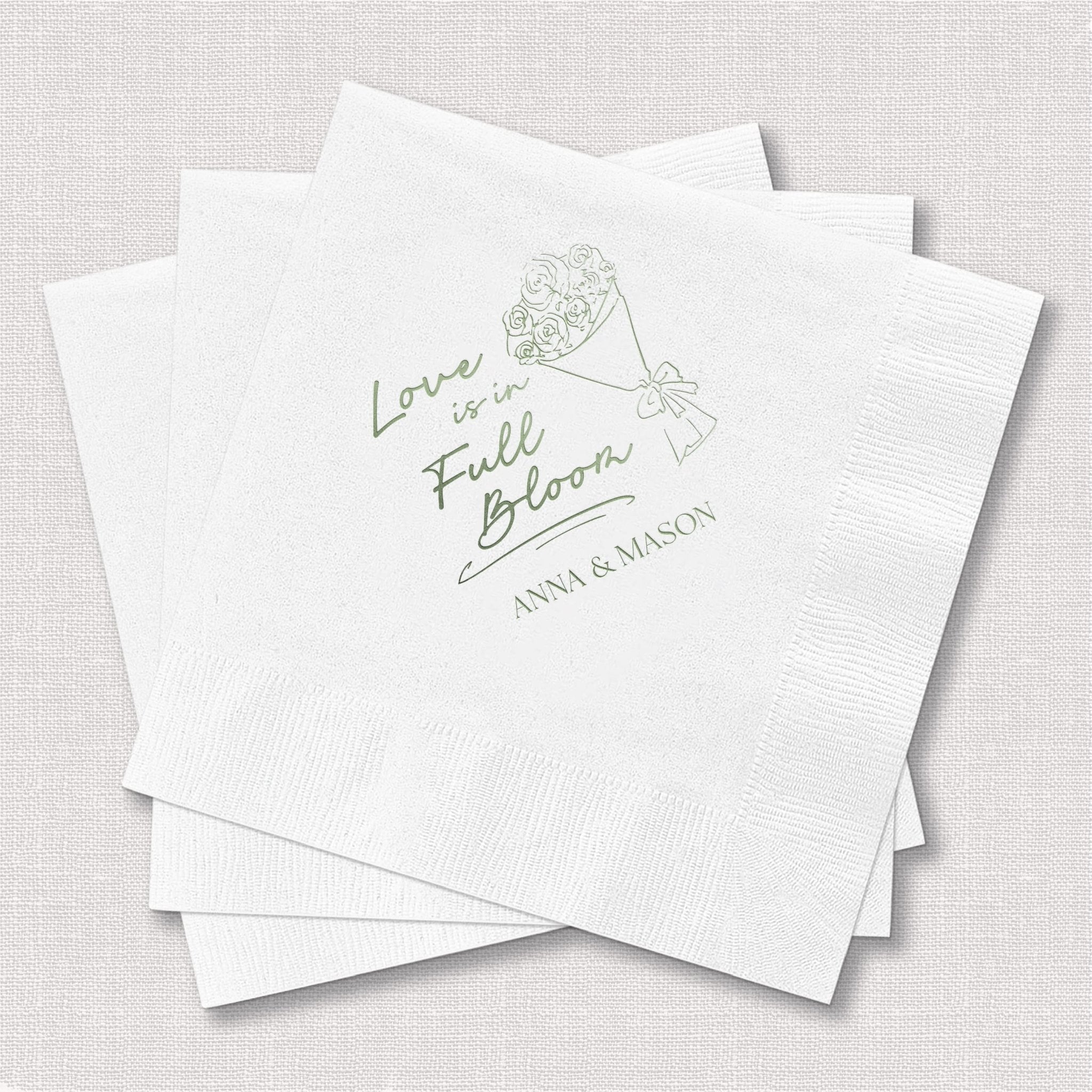 White cocktail napkins with green foil-stamped line drawing of flowers and "Love is in Full Bloom" text with couple's names