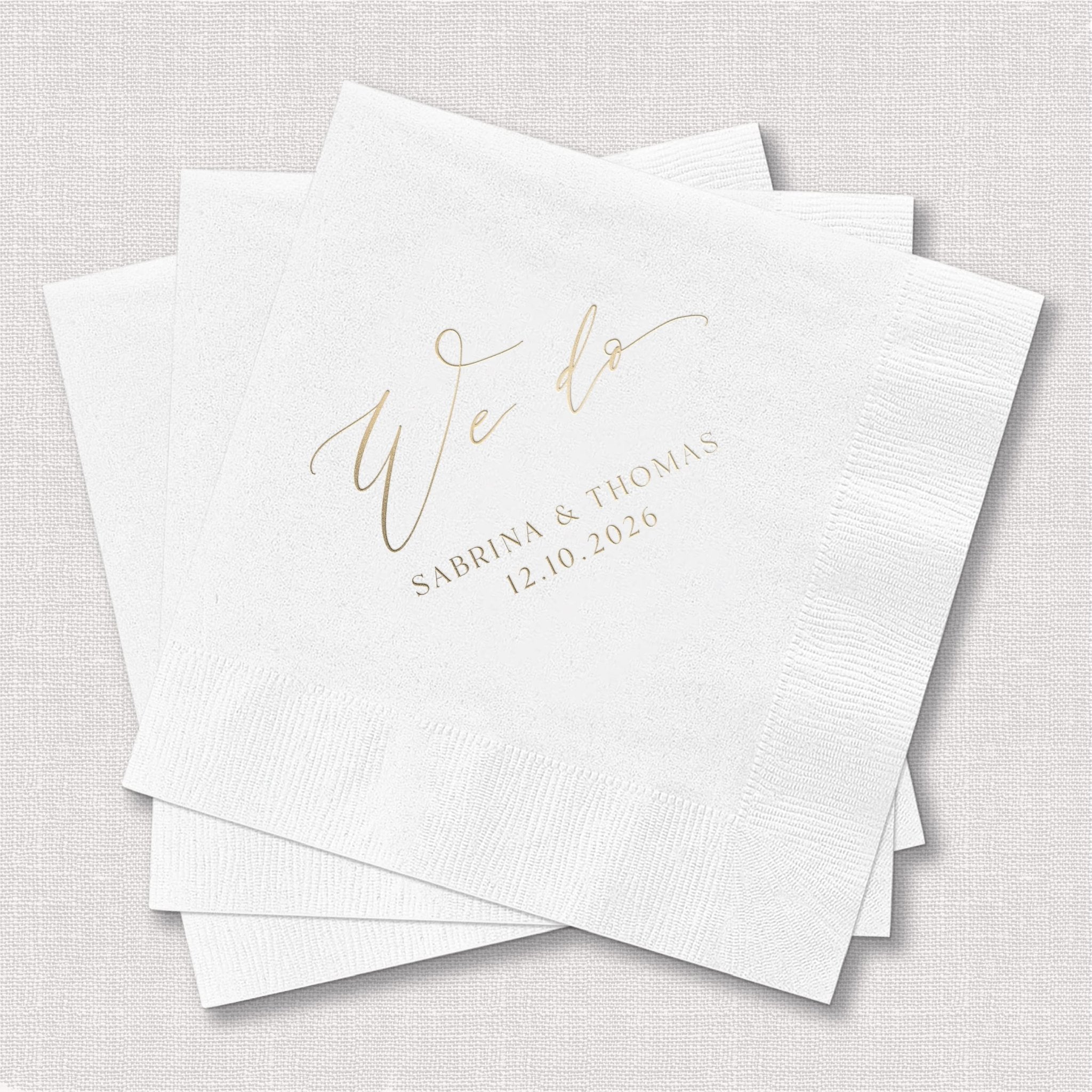 White paper napkins with gold foil stamped "we do" text and couple names with wedding date