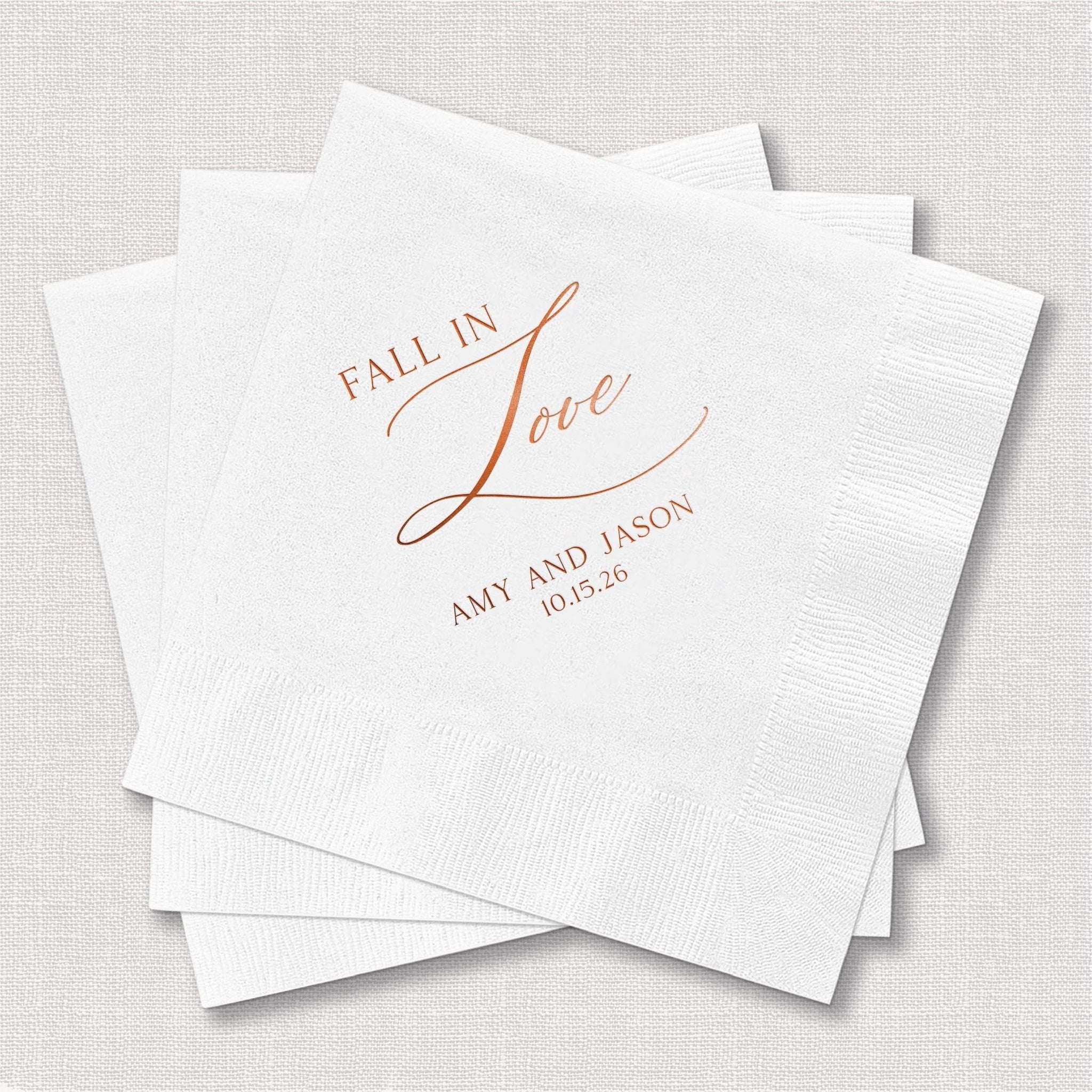White cocktail napkins with rose gold foil stamped text reading "FALL IN LOVE" and personalized names and date below.