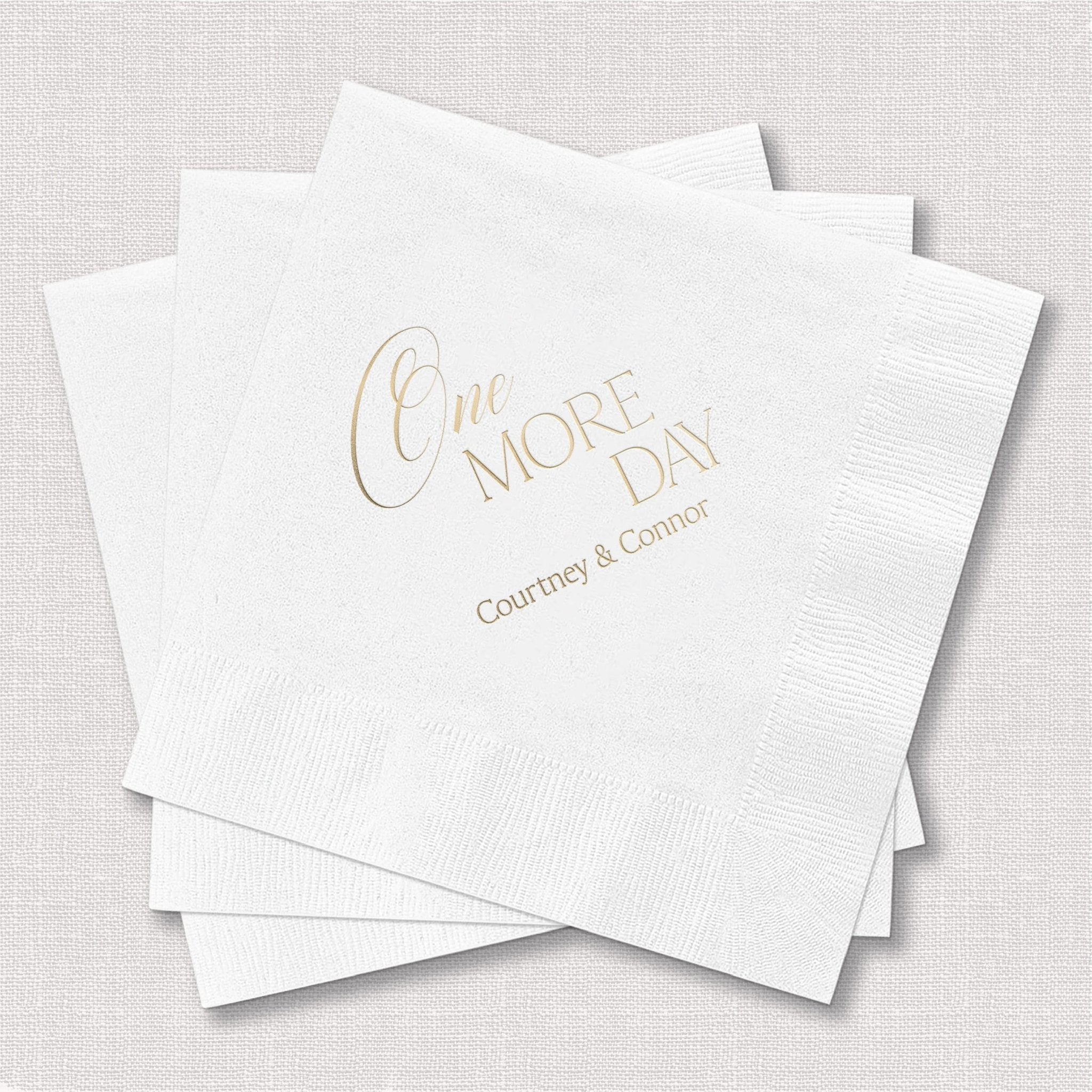 Stack of white cocktail napkins with gold foil stamped text reading "One More Day" and couple's names below.