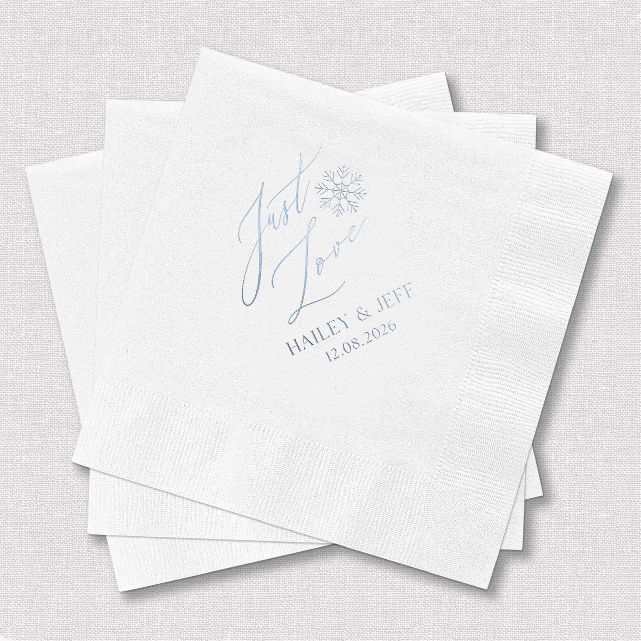White foil-stamped napkins with blue script "Just Love" text, couple's names and wedding date in elegant lettering and floral design.