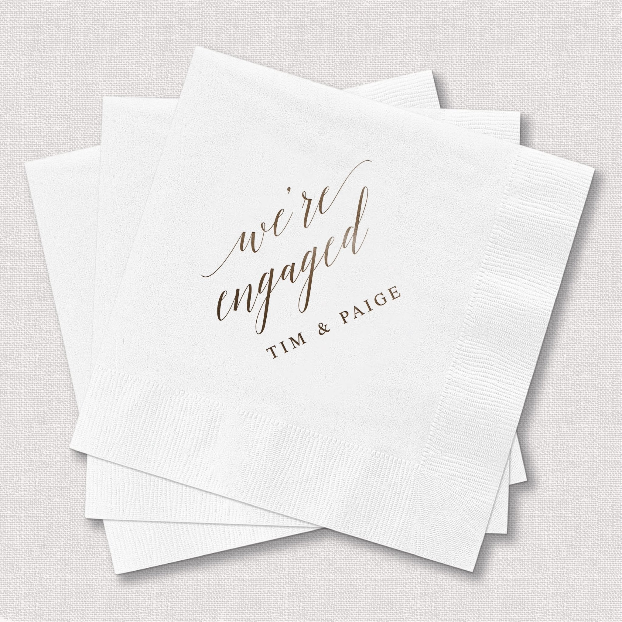 White cocktail napkins with "we're engaged" in script and "TIM & PAIGE" personalized below in elegant foil stamping.