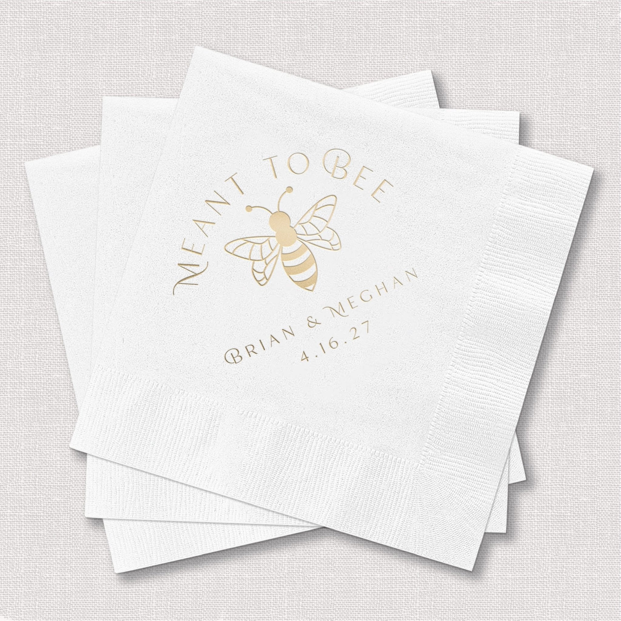 White cocktail napkins with gold foil bee design and "Meant to Bee" text personalized with names and wedding date.
