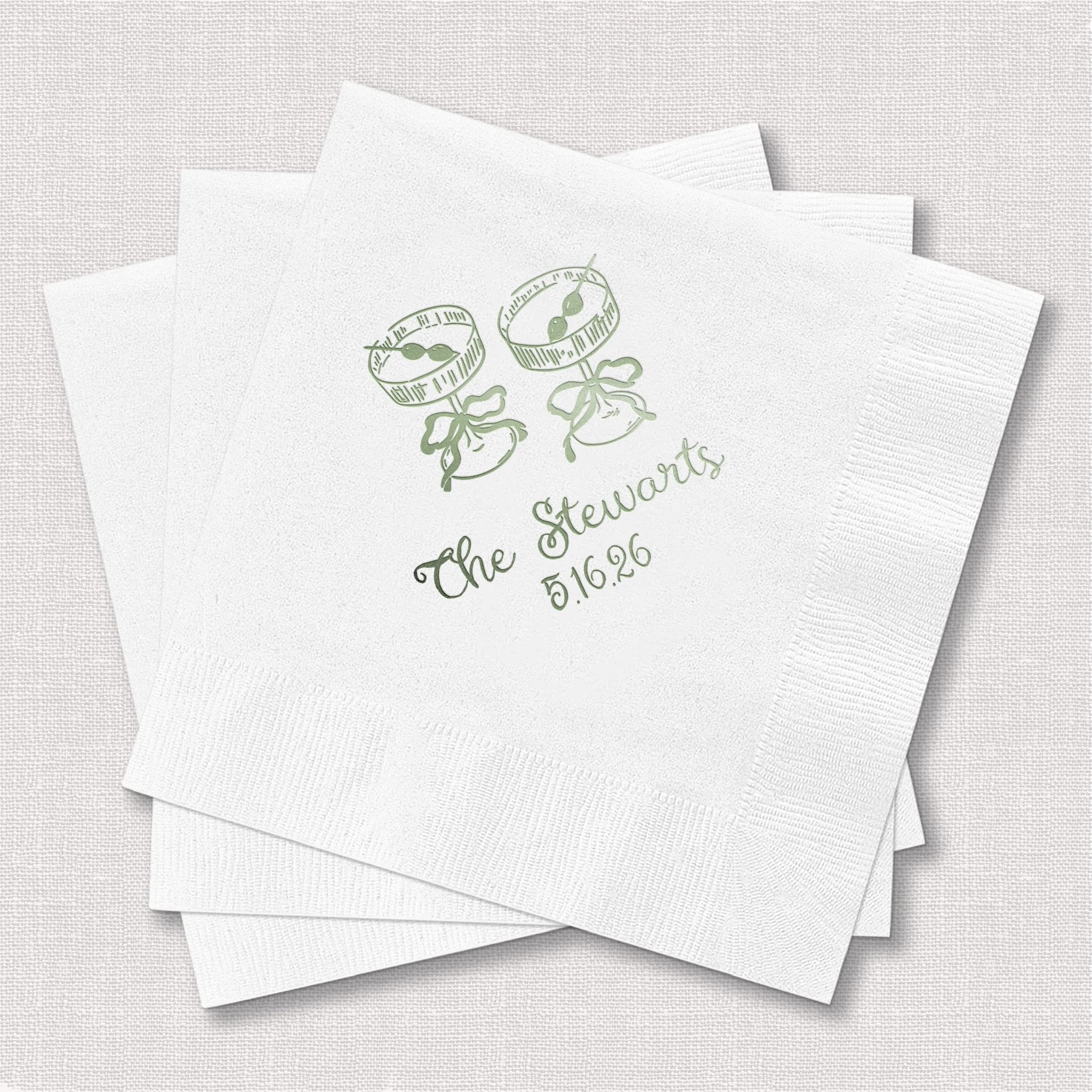 Stack of white cocktail napkins with sage green foil-stamped design featuring two champagne glasses and "The Newlyweds" text with wedding date.