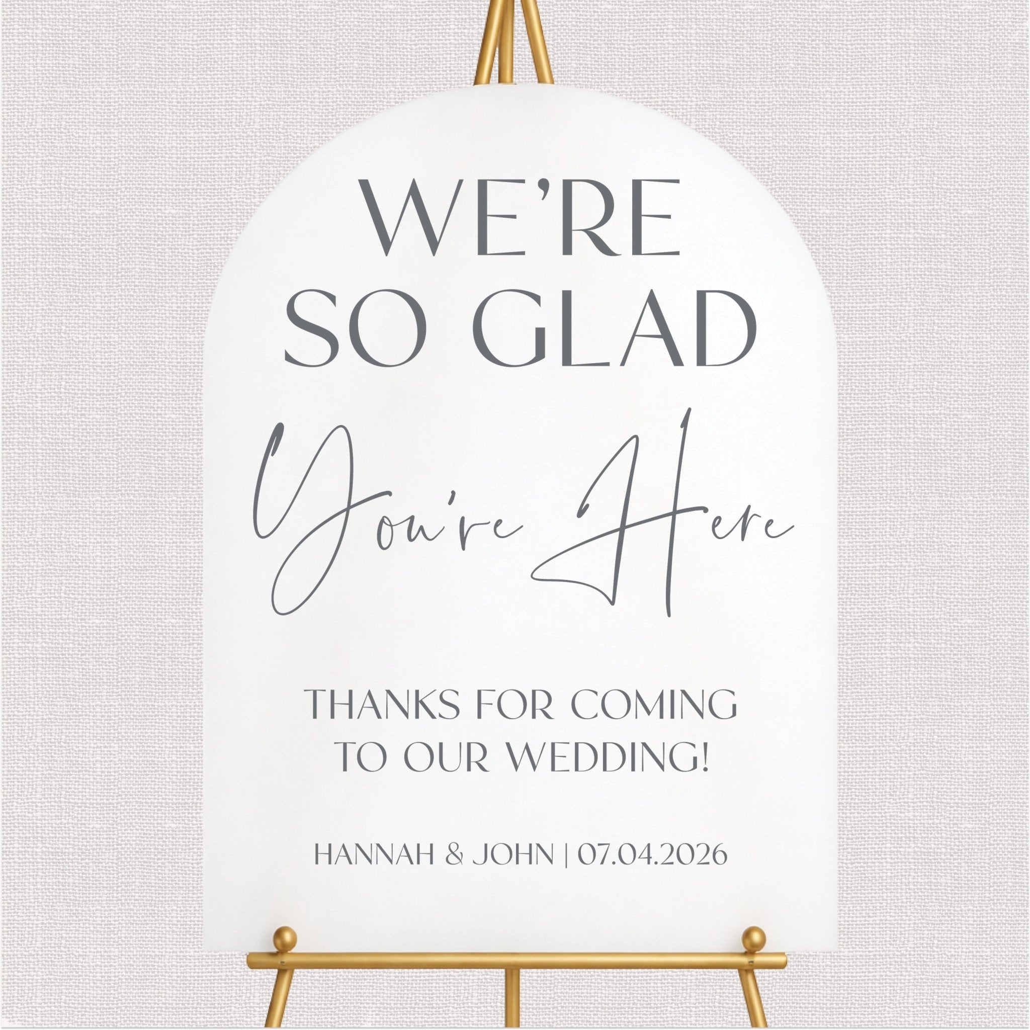 White wedding welcome sign with gray script and serif text, personalized couple names and date, displayed on gold easel.