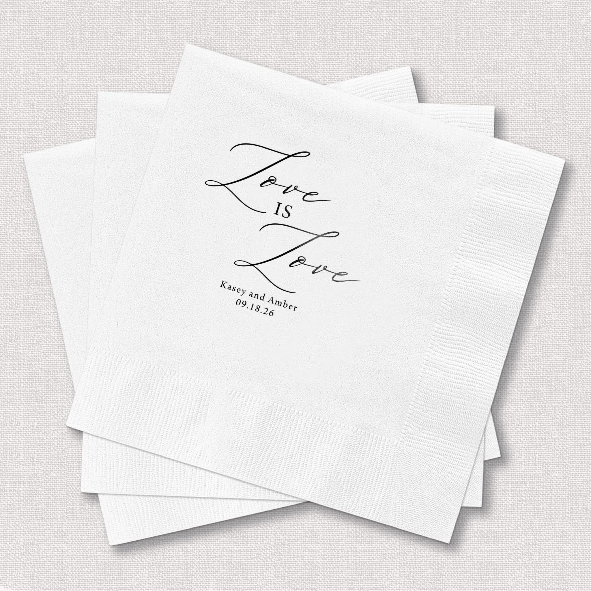 White napkins with "Love is Love" script text and personalized wedding date details in elegant foil stamping.