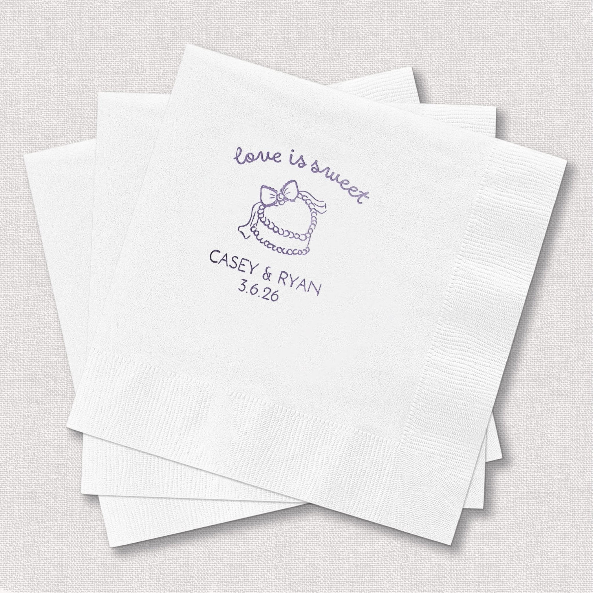 White cocktail napkins with "love is sweet" text, cake illustration, and "Casey & Ryan 5.6.26" personalization in purple foil stamping