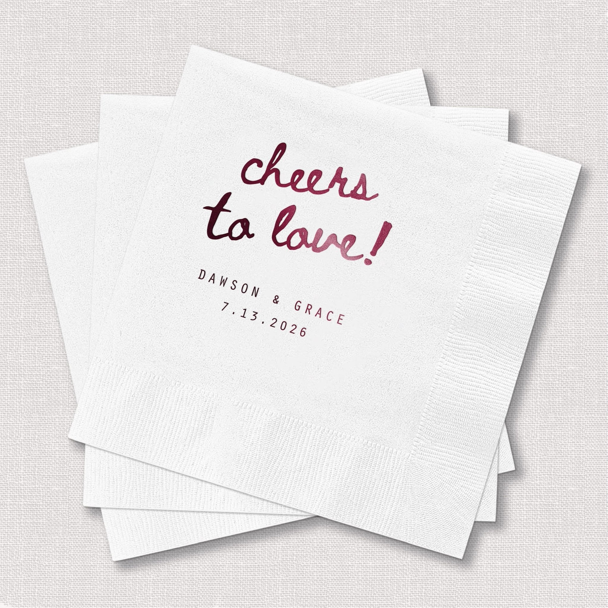 Stack of white beverage napkins with burgundy foil-stamped "cheers to love" text and personalized names and date below.