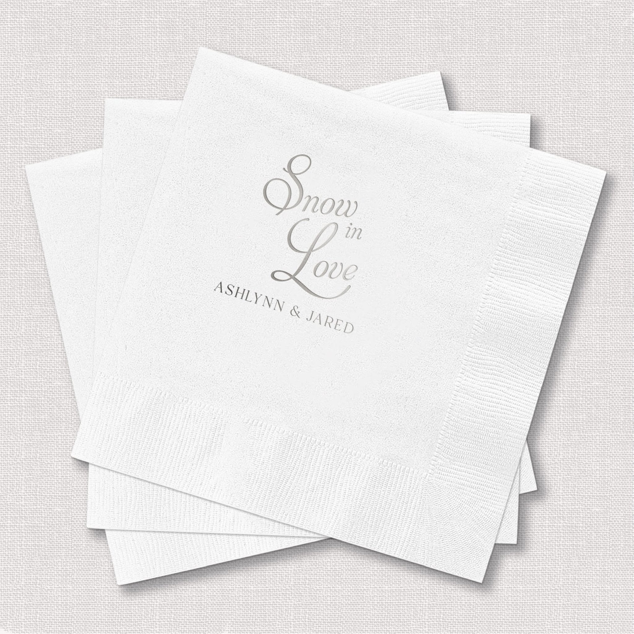 Stack of white napkins with silver foil stamped text reading "Snow in Love" and personalized names for a wedding.