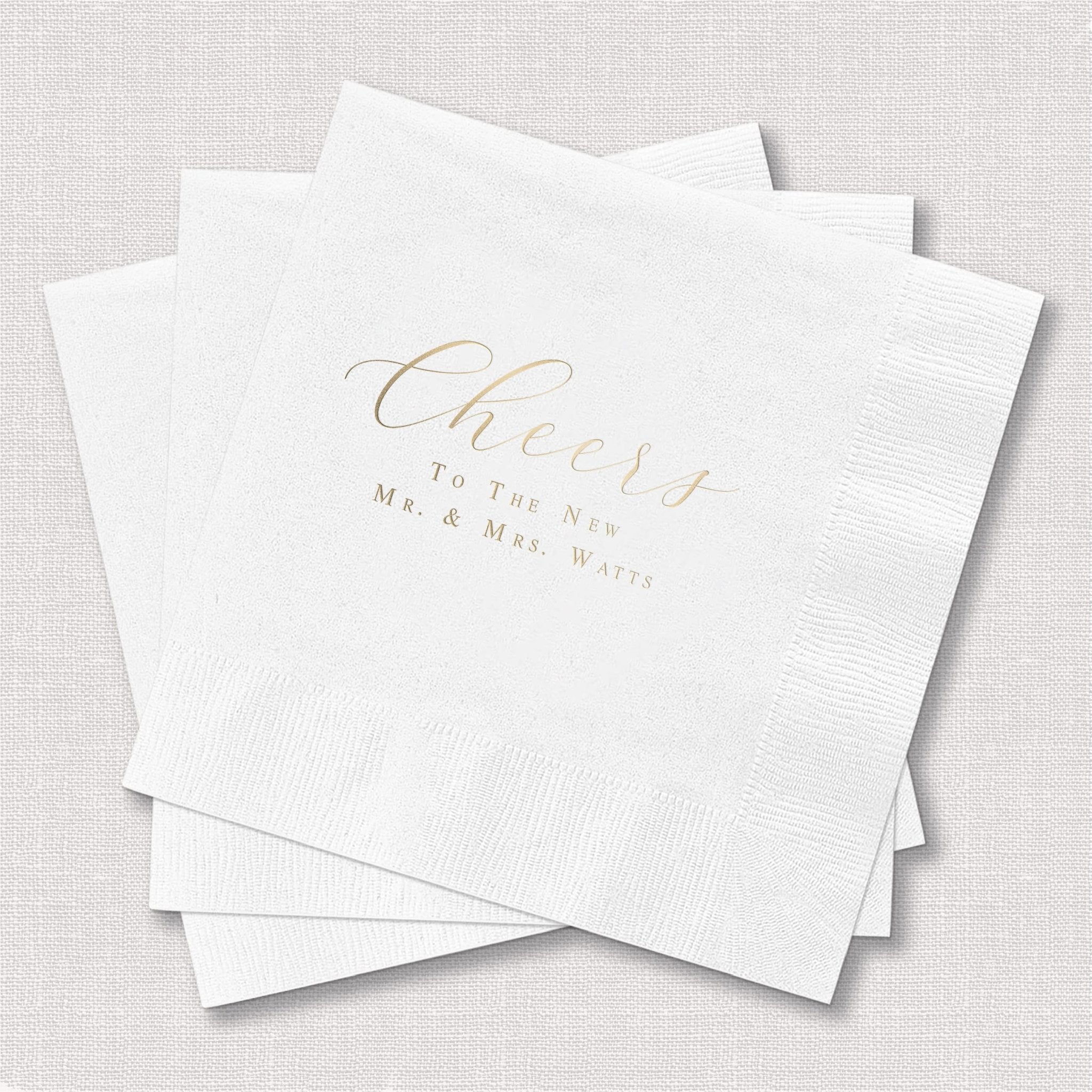 Stack of white napkins with gold foil stamped text reading "Cheers to the Mr. and Mrs. With" for wedding celebration.