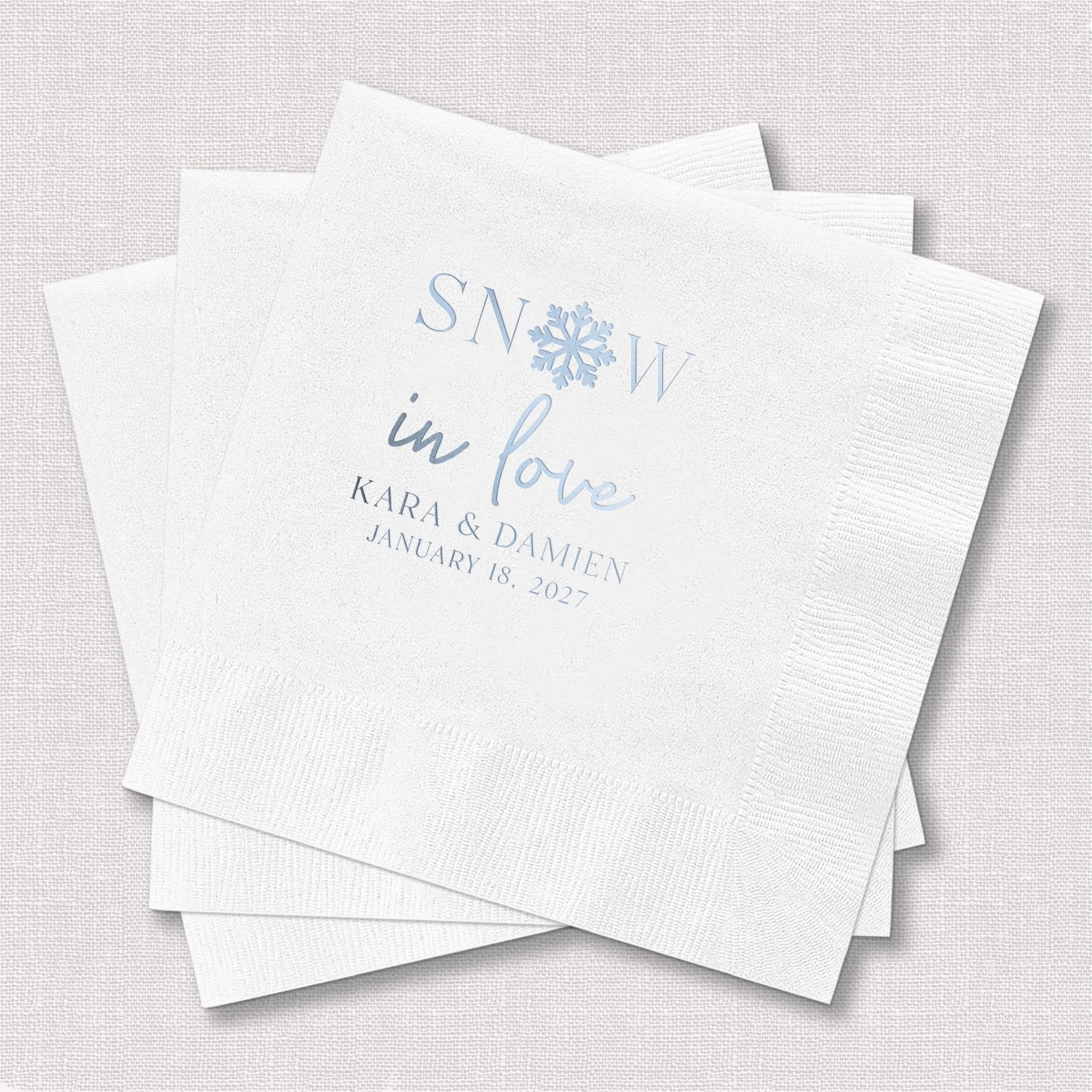 Stack of white napkins with blue foil-stamped text reading "SNOW in love" and personalized names and date below
