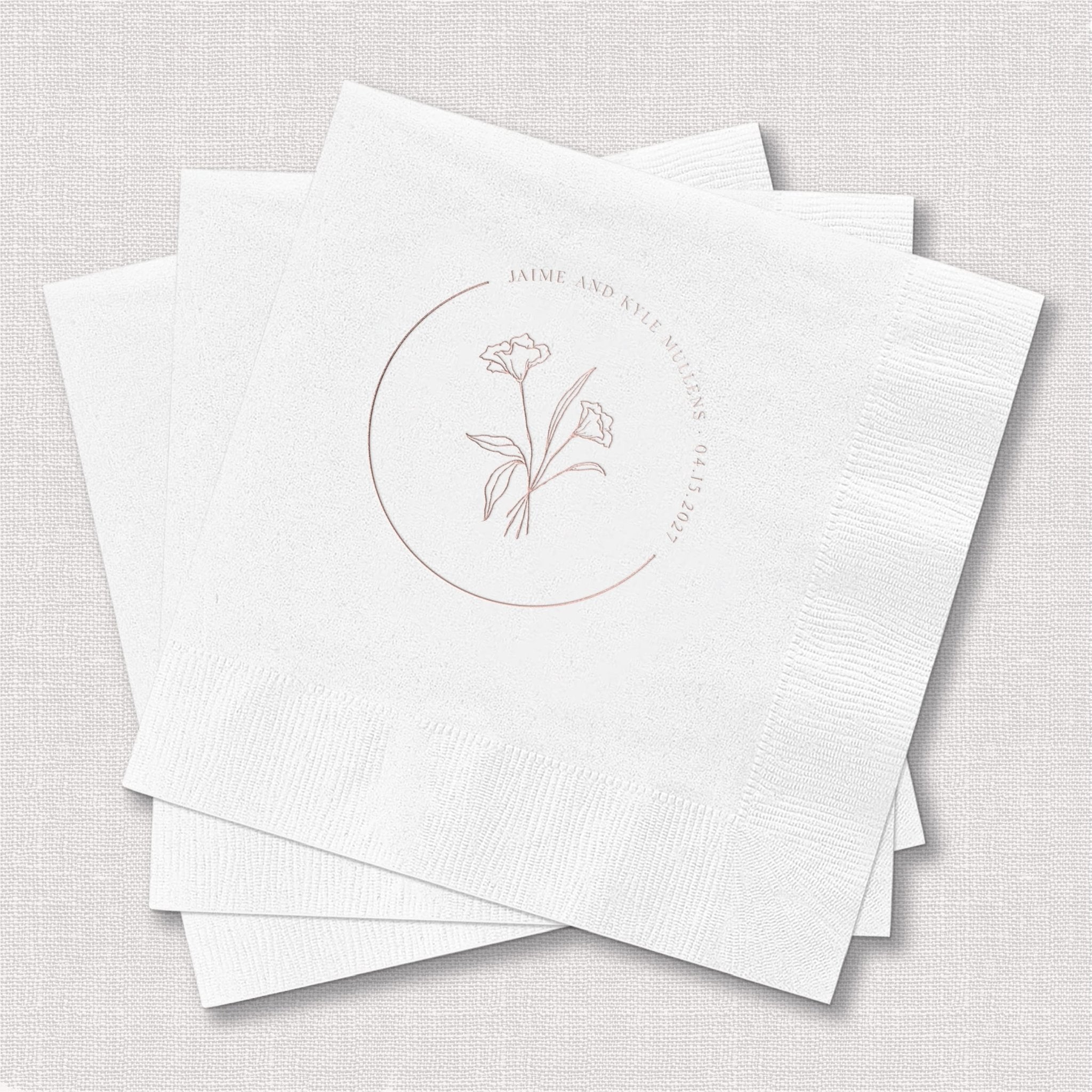 White cocktail napkins with delicate floral design and circular text in rose gold foil stamping