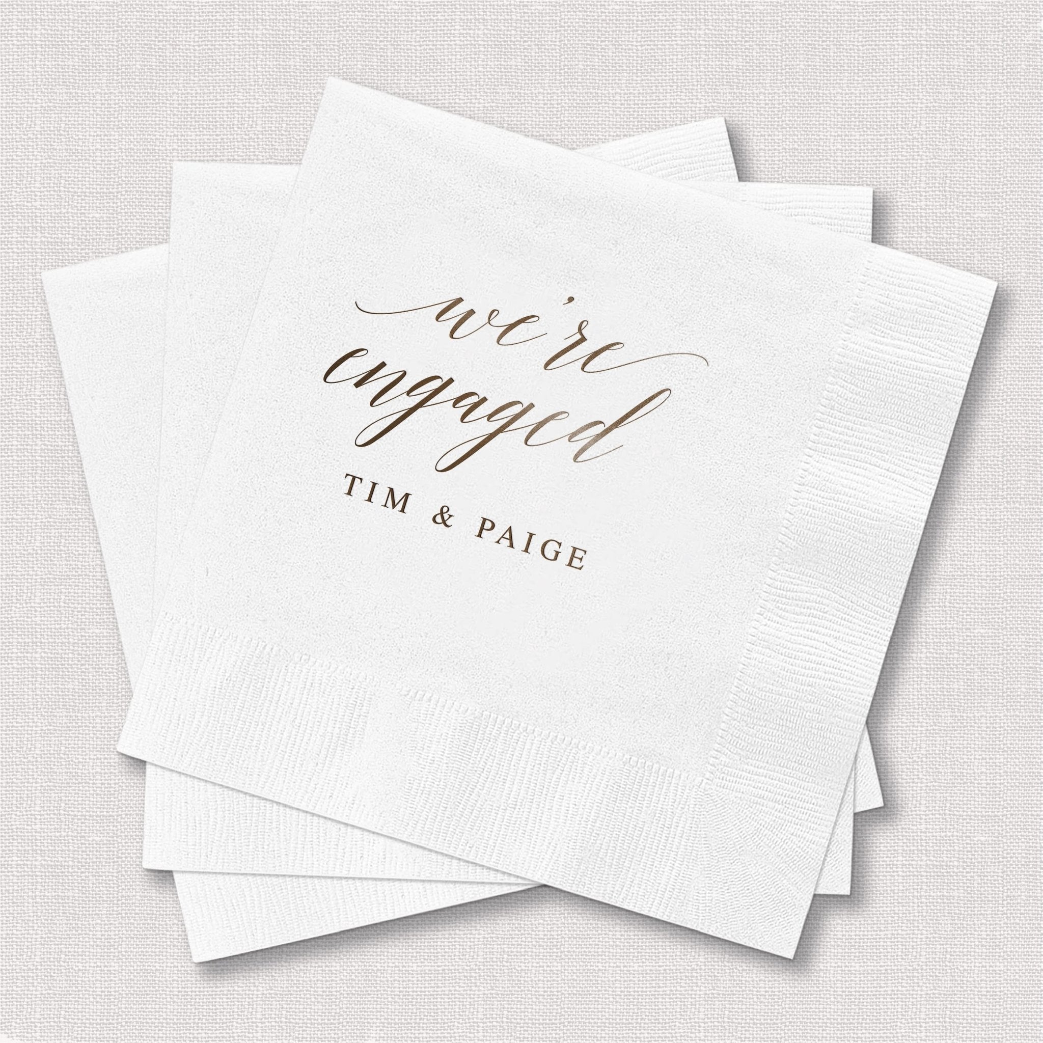 Stack of white cocktail napkins with gold foil text reading "we're engaged" and personalized names "Tim & Paige" below.