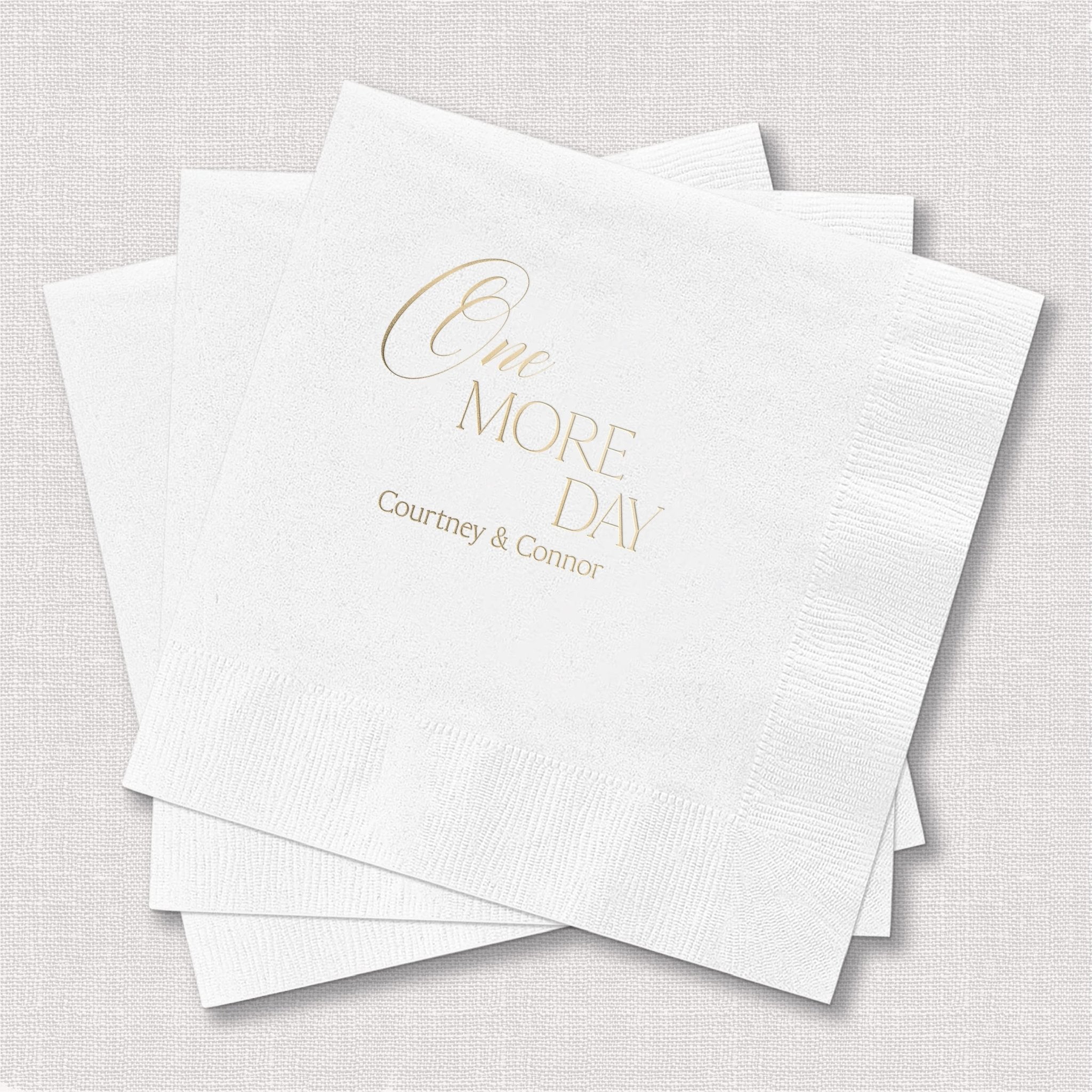 White cocktail napkins with gold foil stamped text reading "One MORE DAY" and personalized couple names below.