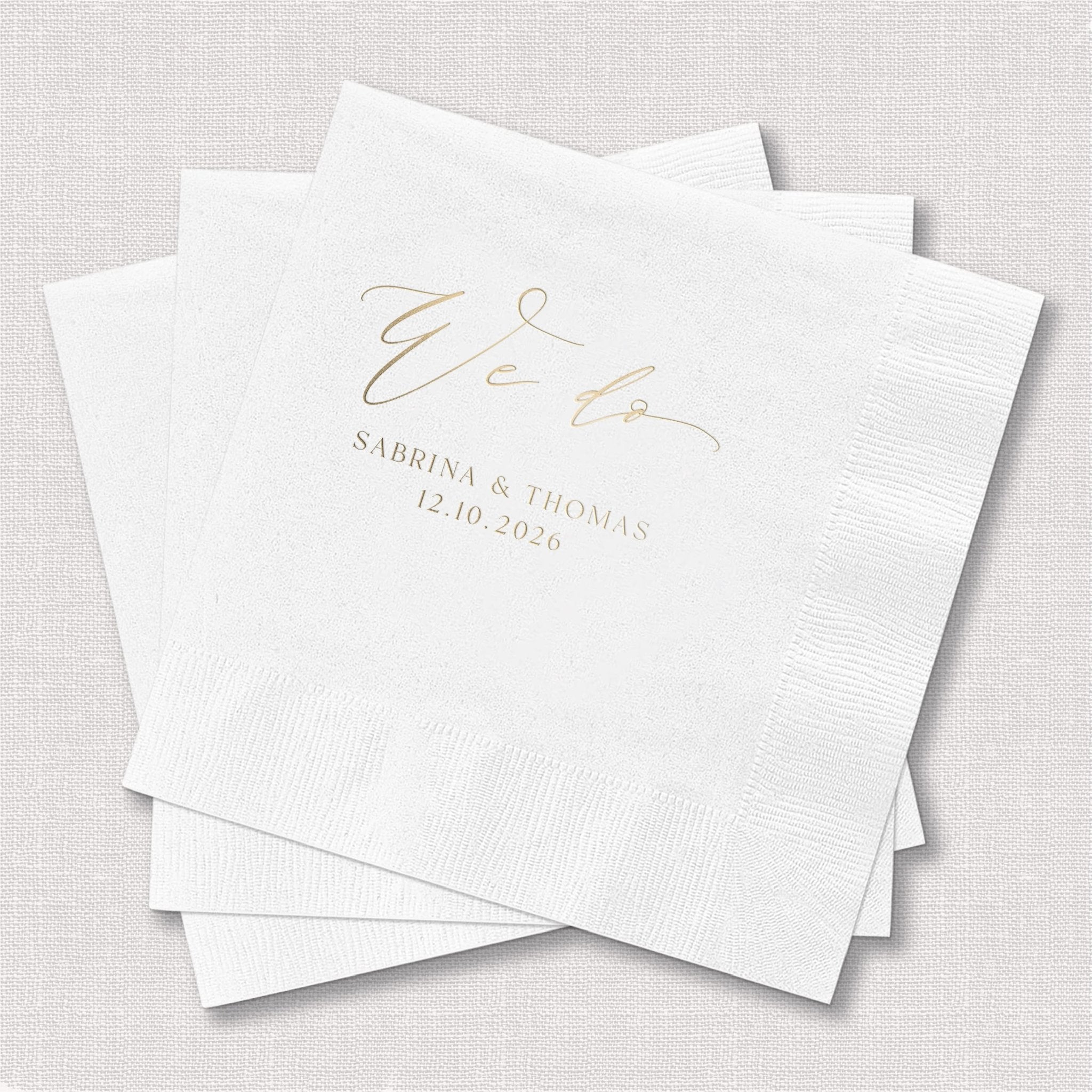 White foil stamped napkins with gold "We Do" script and couple's names Sabrina & Thomas with wedding date 13.10.2025