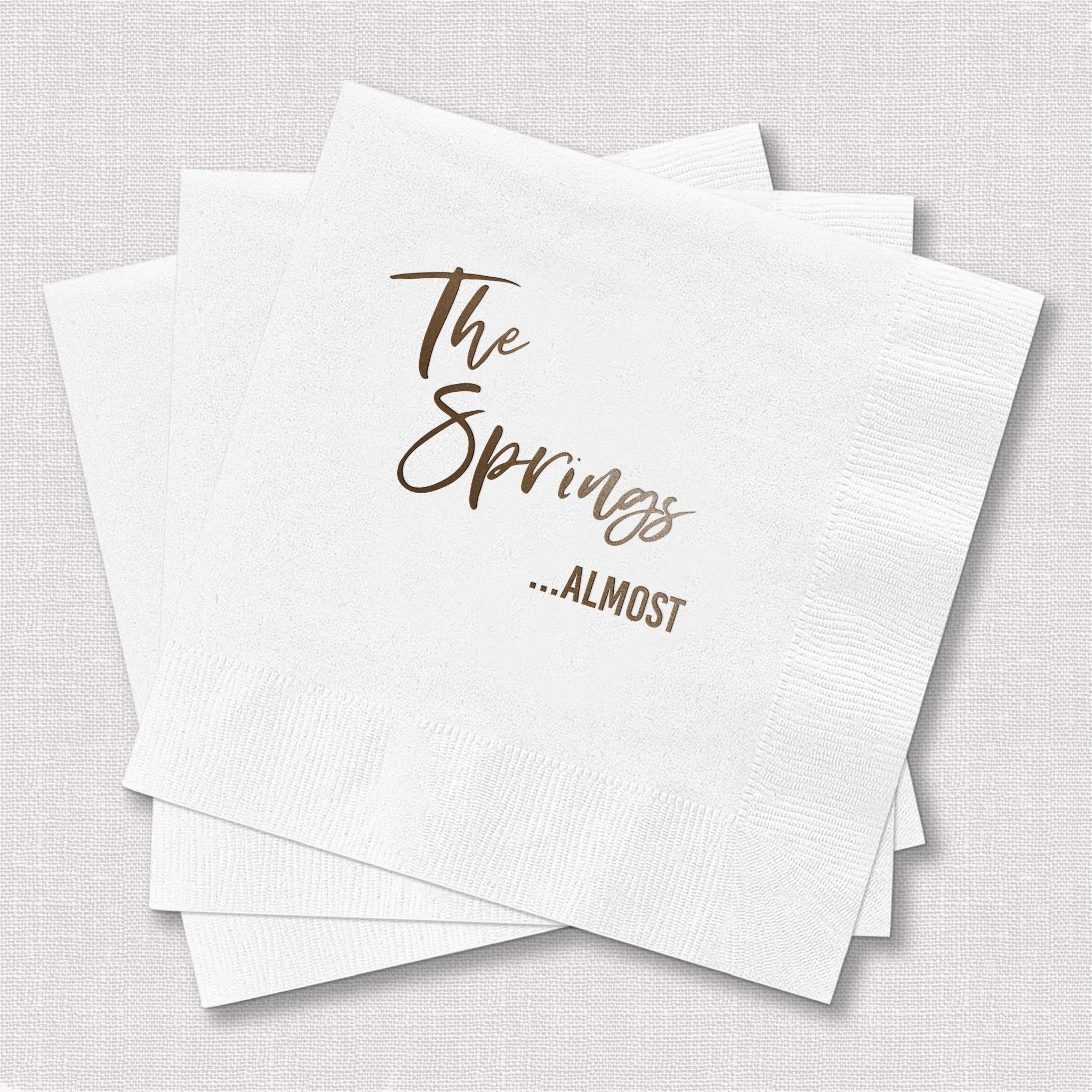 White cocktail napkins with gold foil text reading "The Springs ...ALMOST"