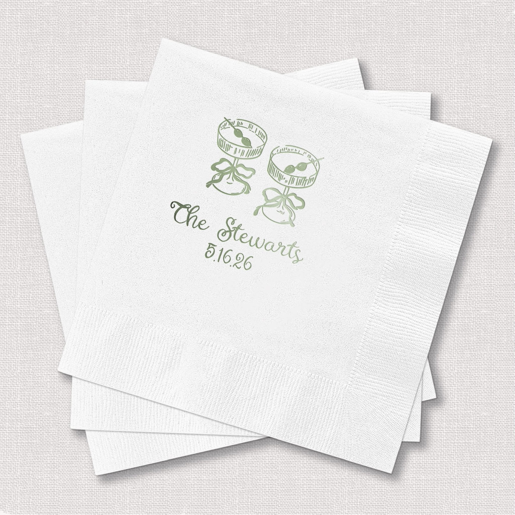 White cocktail napkins with sage green foil-stamped champagne glasses and "The Stewarts 8.16.25" text