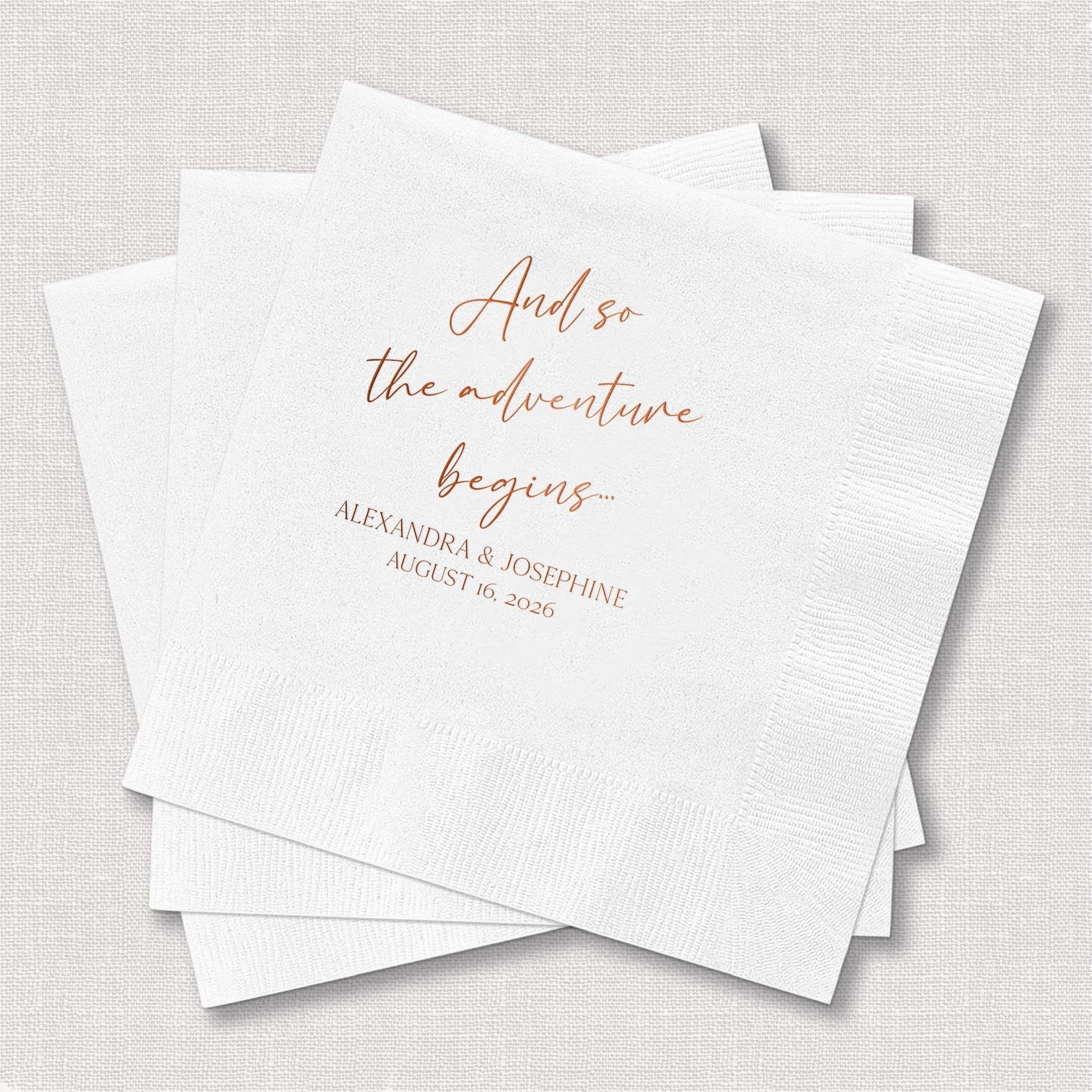 White paper napkins with rose gold foil-stamped text reading "And so the adventure begins" and couple names with wedding date.