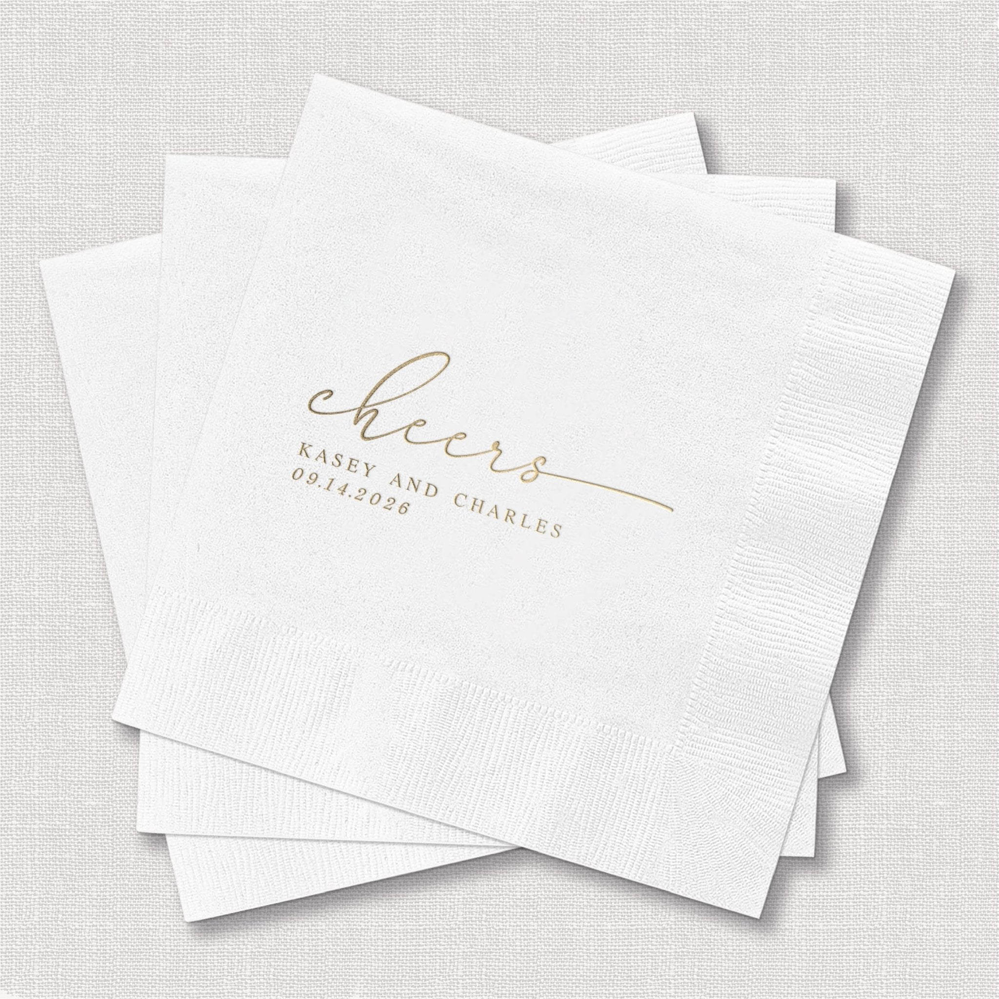 Stack of white beverage napkins with gold foil stamped "cheers" script and personalized names and date below.