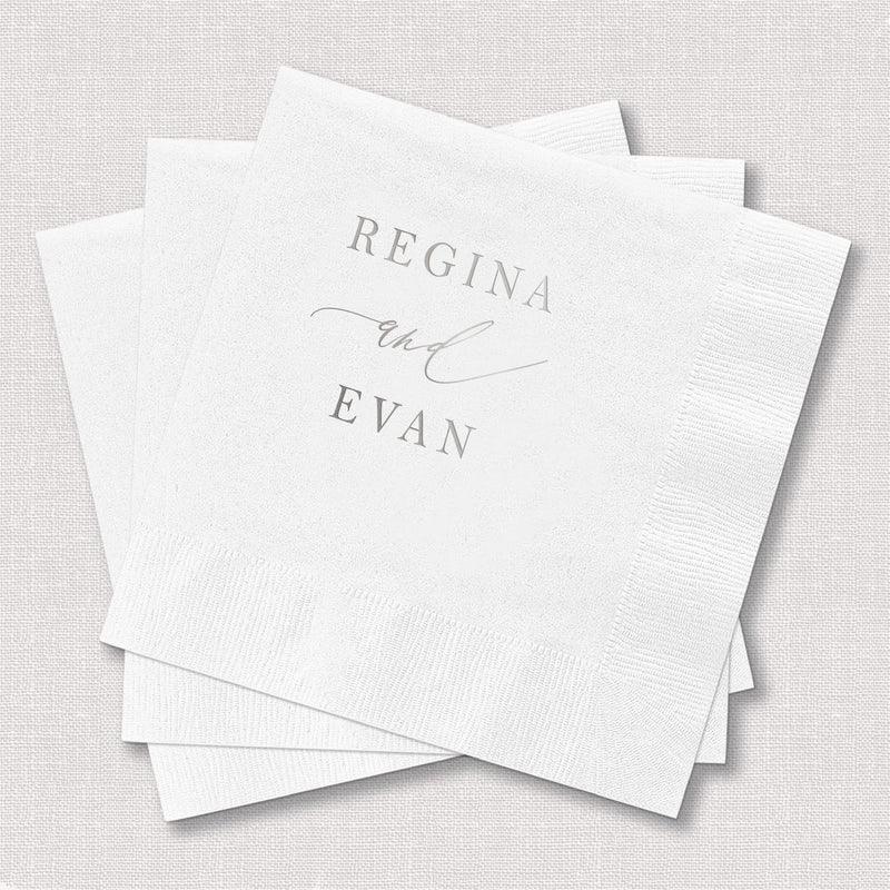 Stack of white cocktail napkins with silver foil-stamped names Regina and Evan in elegant script font