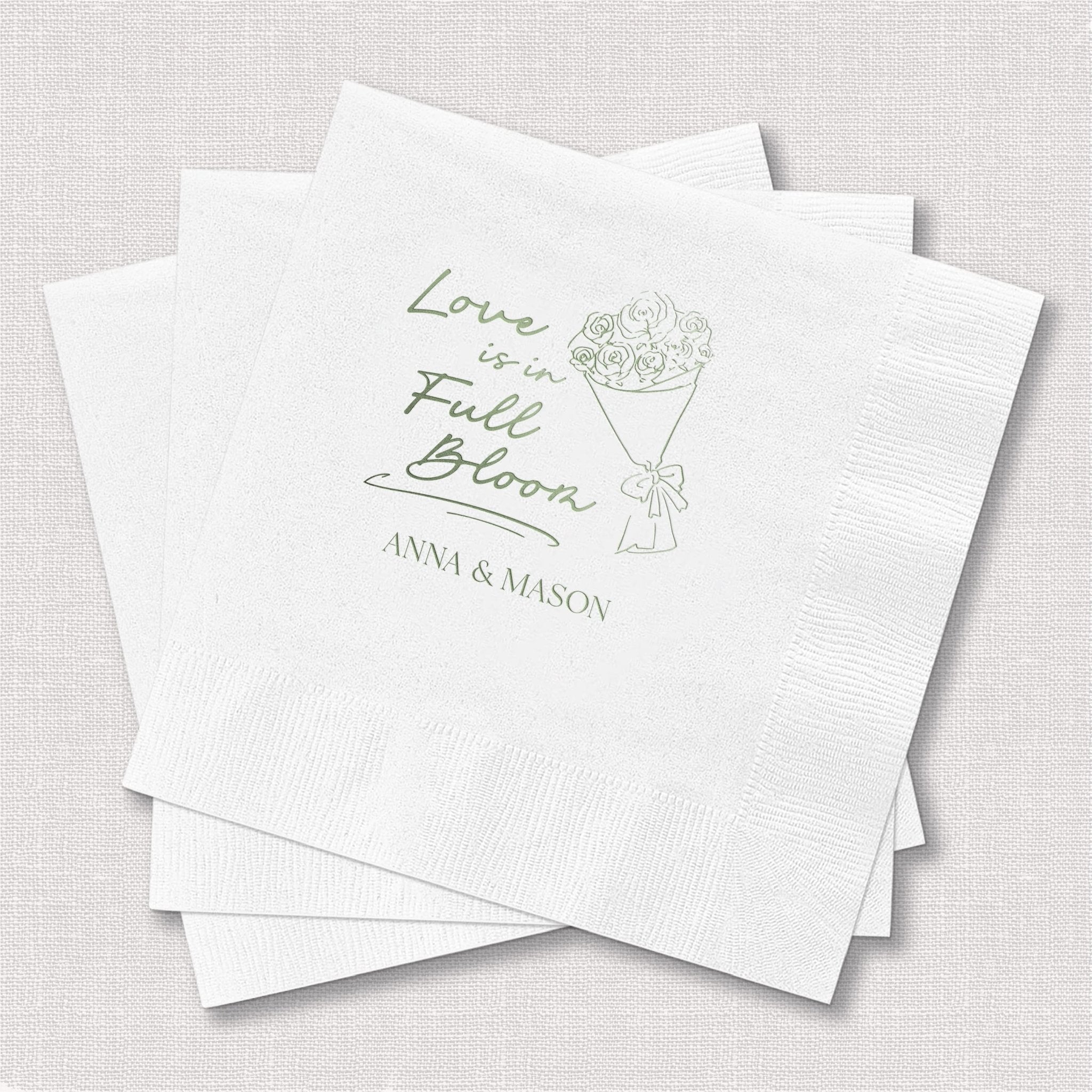 Stack of white napkins with foil-stamped design featuring "Love Is In Full Bloom" text, floral bouquet illustration, and couples names Anna and Mason.