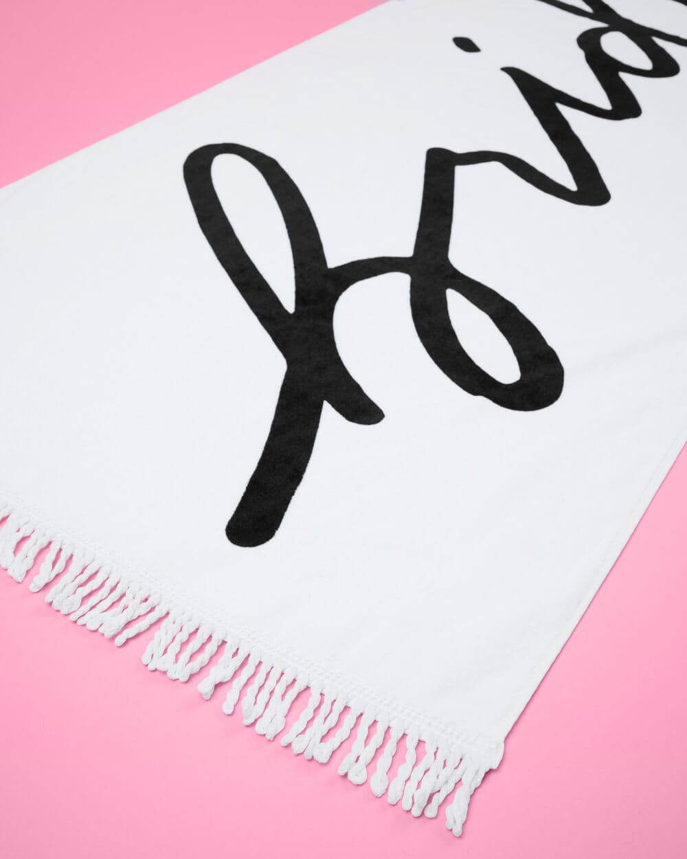 White cotton beach towel with black handwritten-style text and fringe trim on pink background