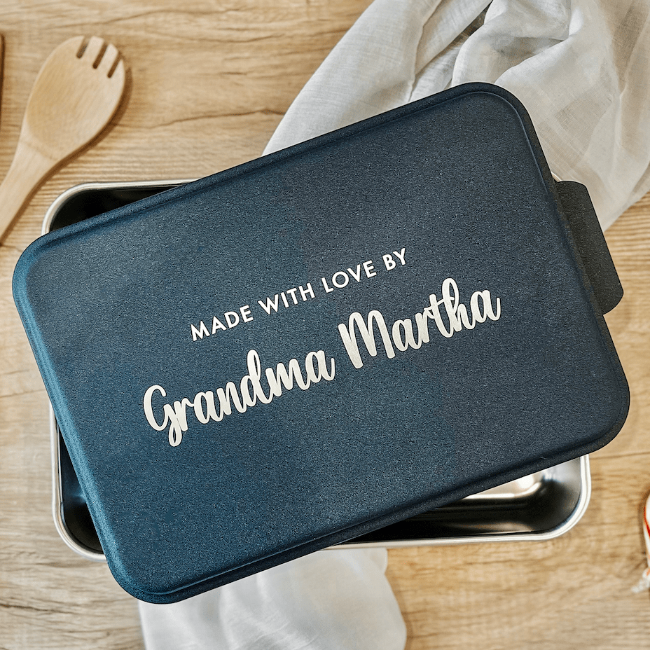 Dark teal rectangular cake pan with "Made with love by Grandma Martha" in white script text on lid, displayed on wooden surface with kitchen linens.