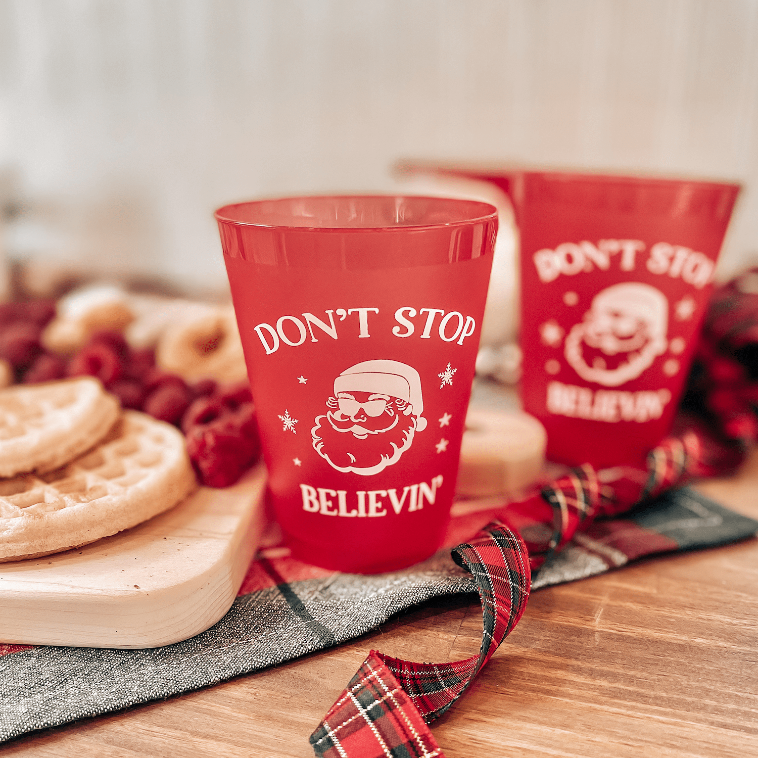 Red frosted shatterproof cups with Santa and "Don't Stop Believin'" text, displayed on holiday table setting with cookies and plaid ribbon.