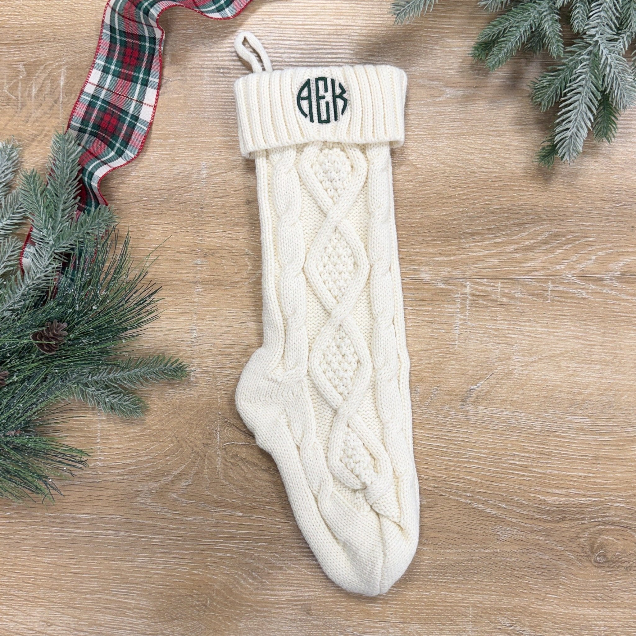 Cream cable knit Christmas stocking with monogram embroidery on white cuff, displayed on wood surface with greenery and plaid ribbon.