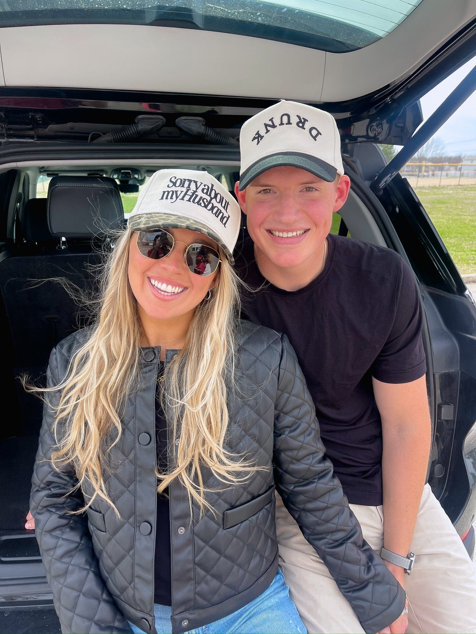 Couple wearing white vintage trucker hats with "Sorry about my Husband" and "Sorry about my Wife" text inside a vehicle.