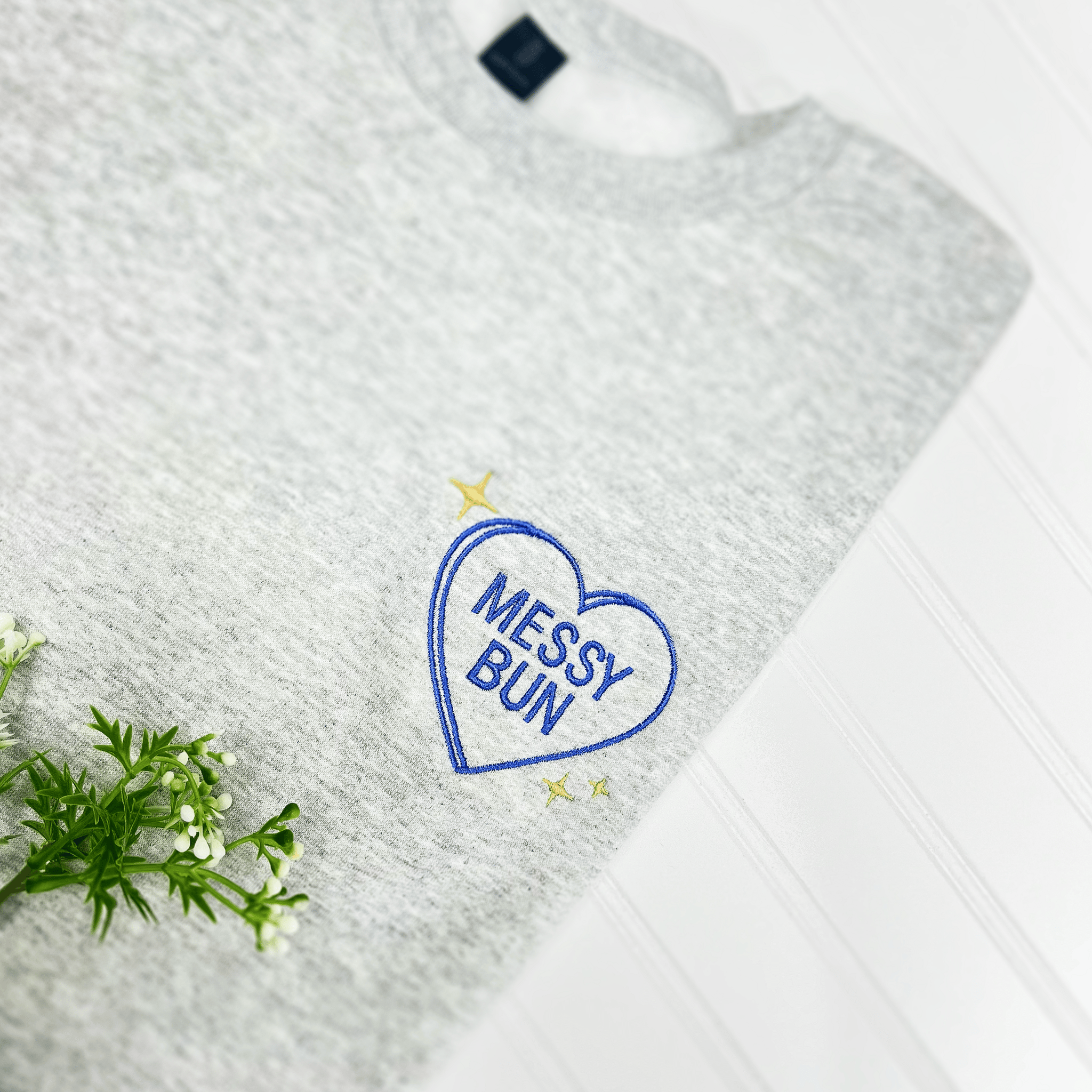 Heather gray sweatshirt with blue embroidered heart design reading "Messy Bun" and small gold star accents.