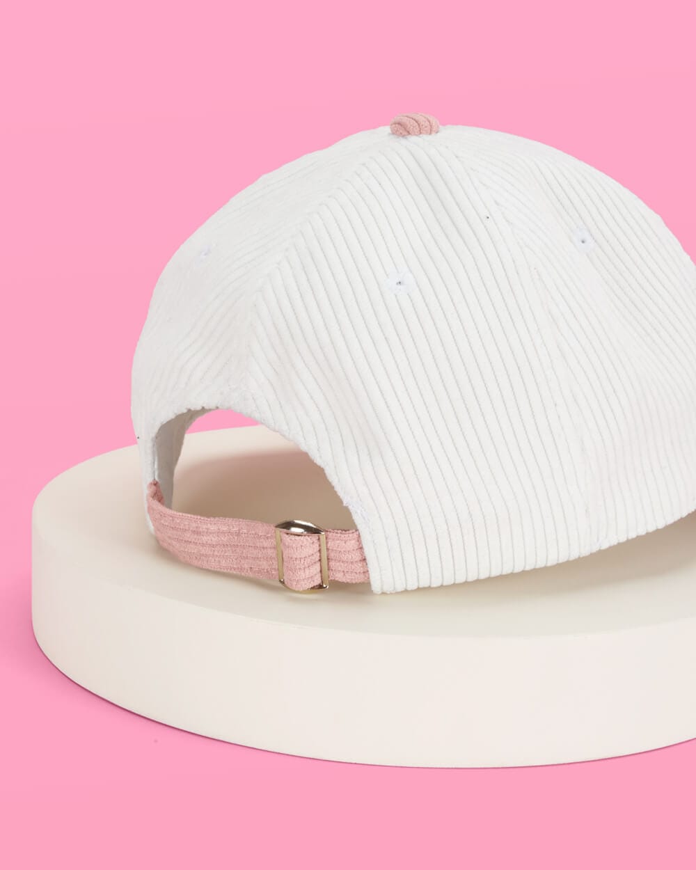 White ribbed baseball cap with pink corduroy back strap and buckle closure on pink background