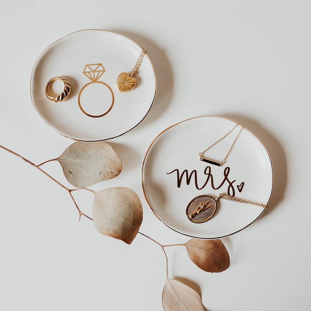 White ceramic ring dish with "mrs" text and gold pendant necklace displayed on surface