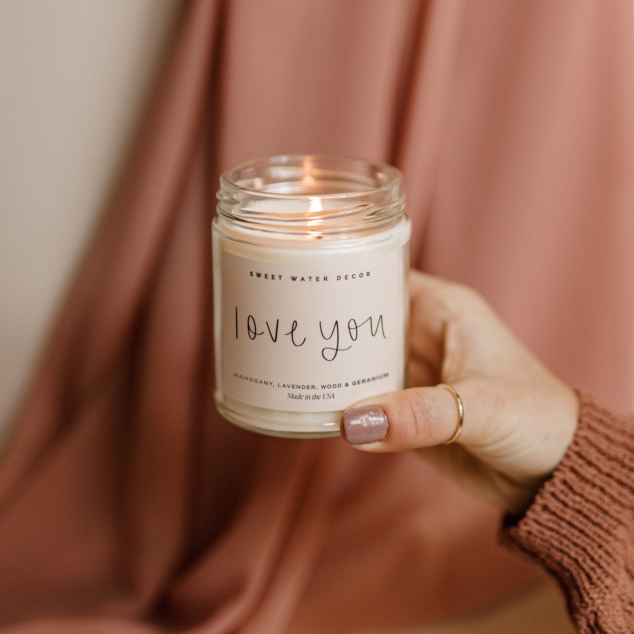 Hand holding clear glass jar candle with "love you" text on cream label against pink fabric background