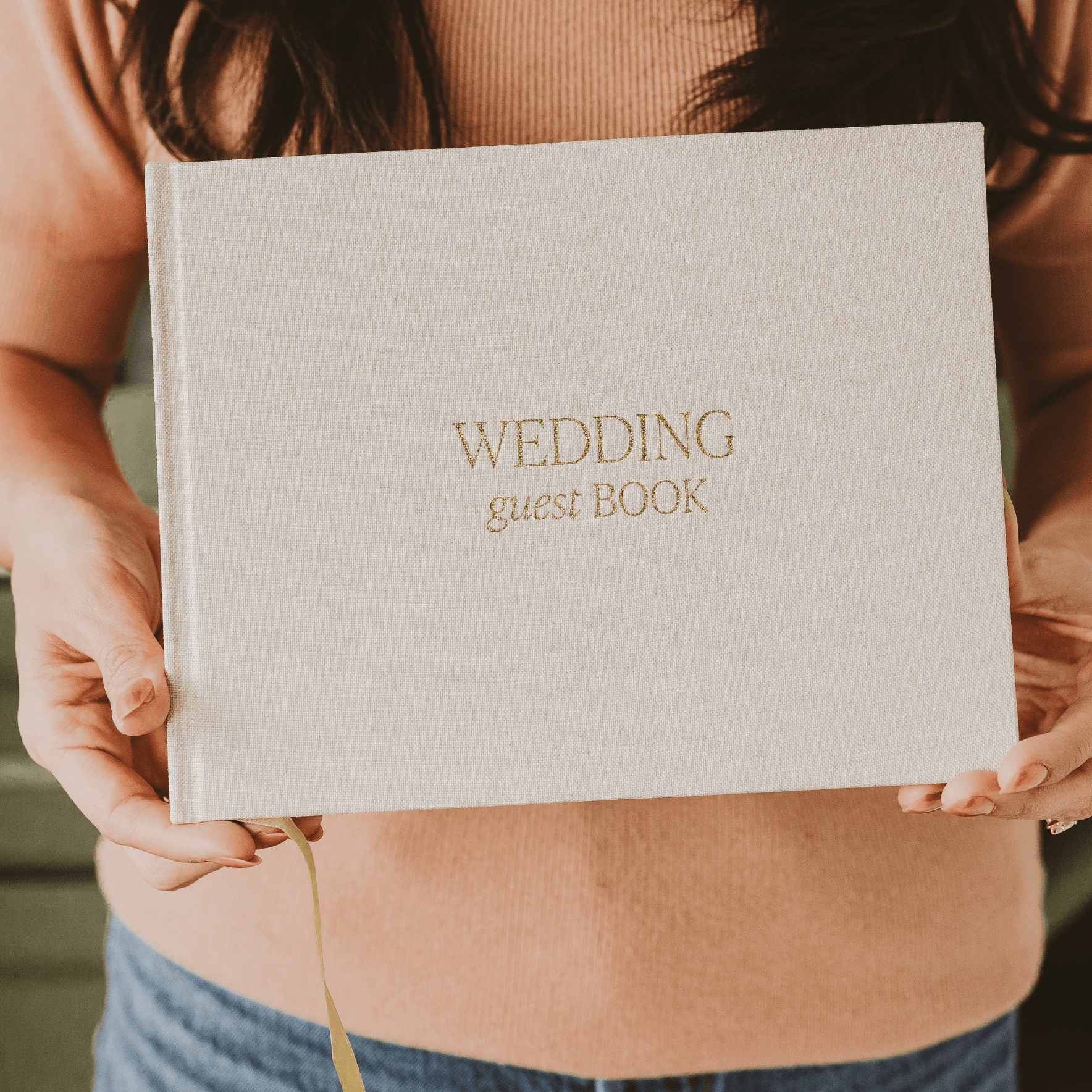 Cream wedding guest book with gold foil text held by woman in tan sweater