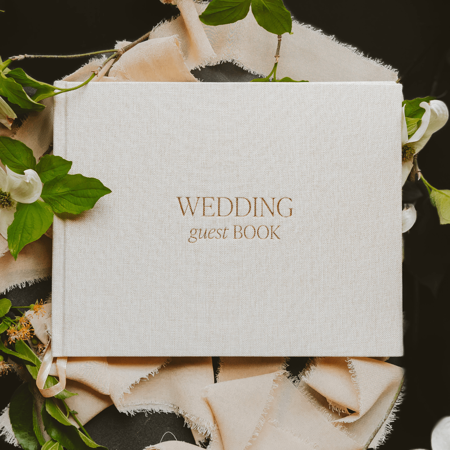 Tan linen wedding guestbook with gold foil text, surrounded by white flowers and greenery