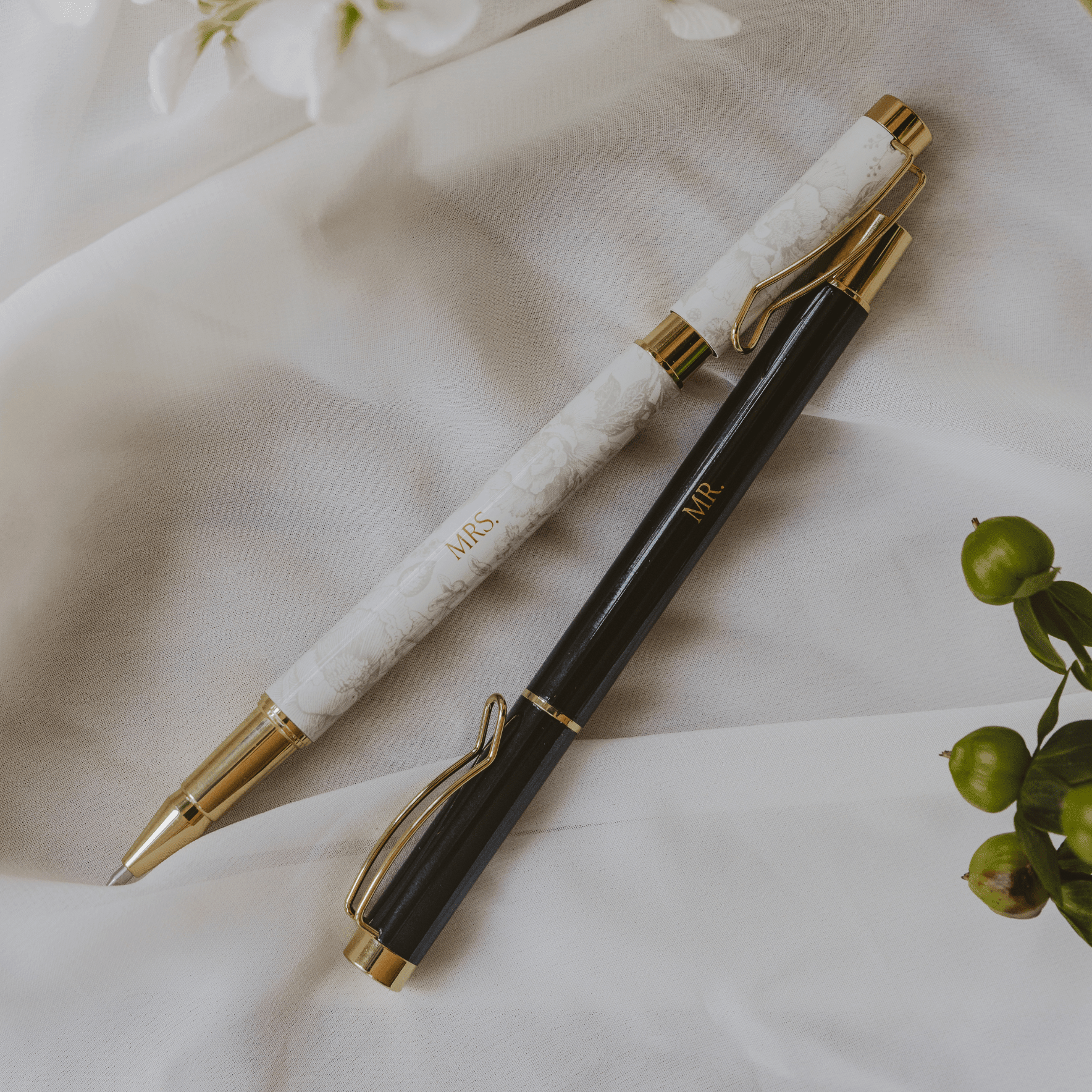 White marble and black ballpoint pens with gold accents, displayed on cream fabric with green berries and white flowers.