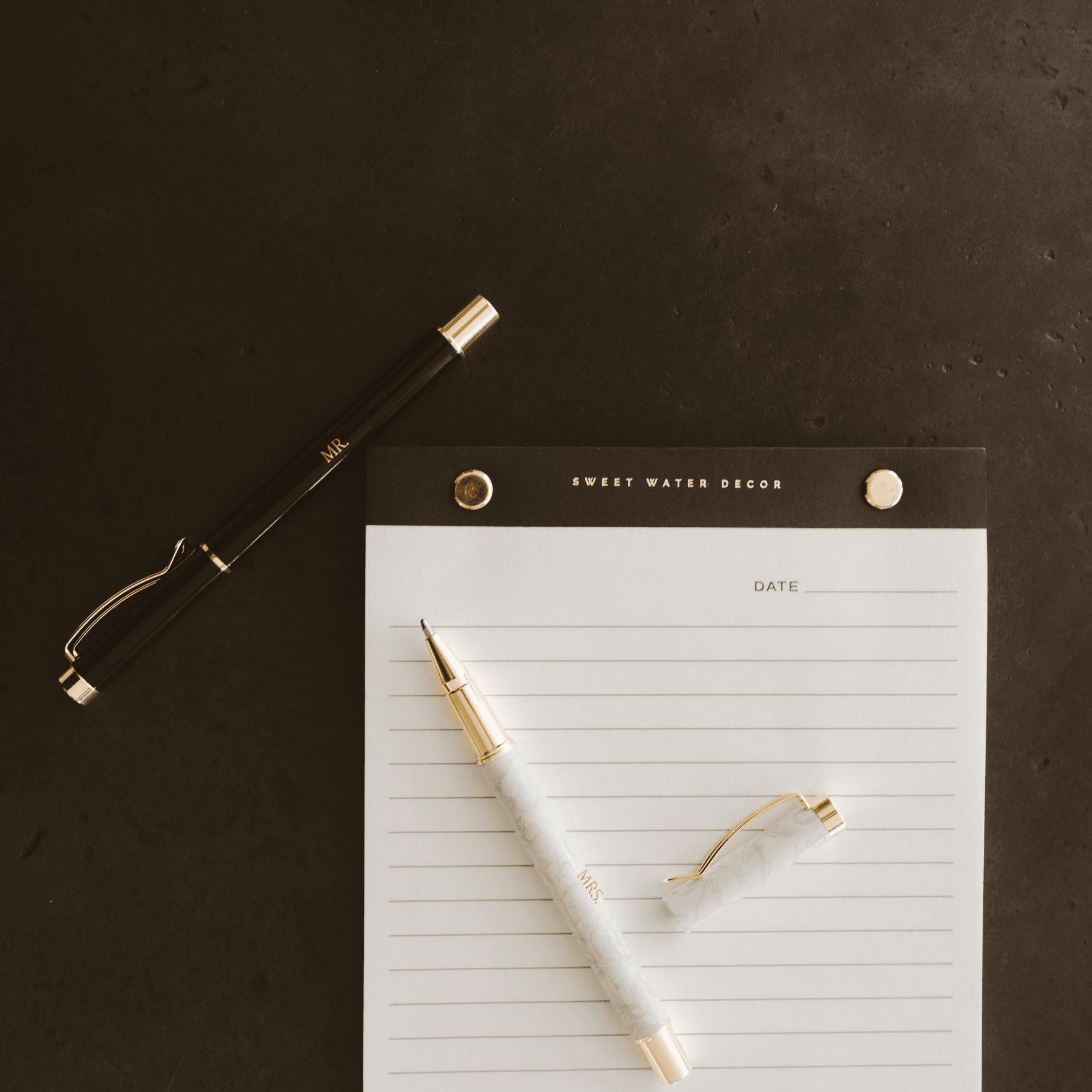 White and gold wedding pen set with lined notepad and decorative accessories on dark background