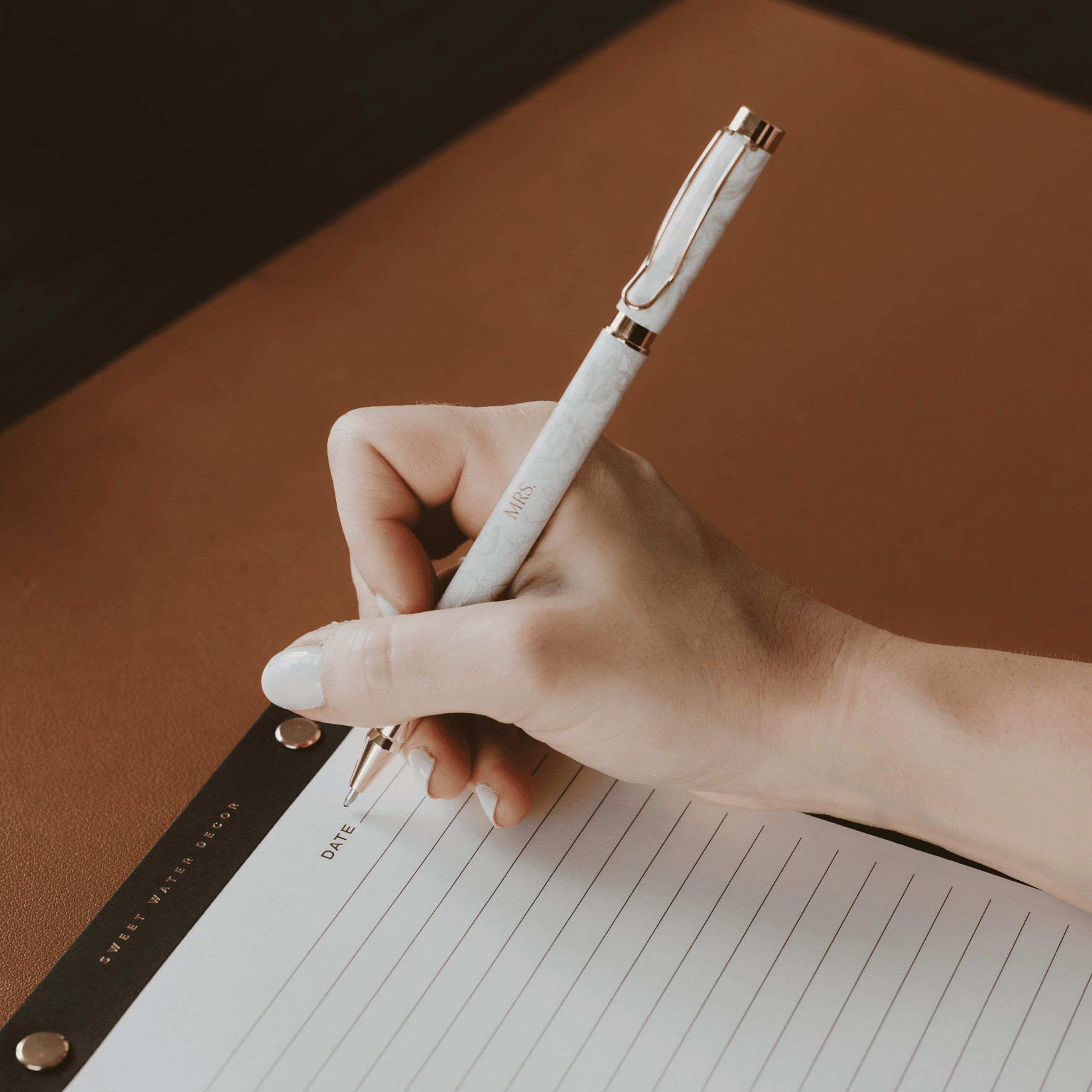 White pen held above lined notebook with black leather organizer visible, styled flat lay on tan background