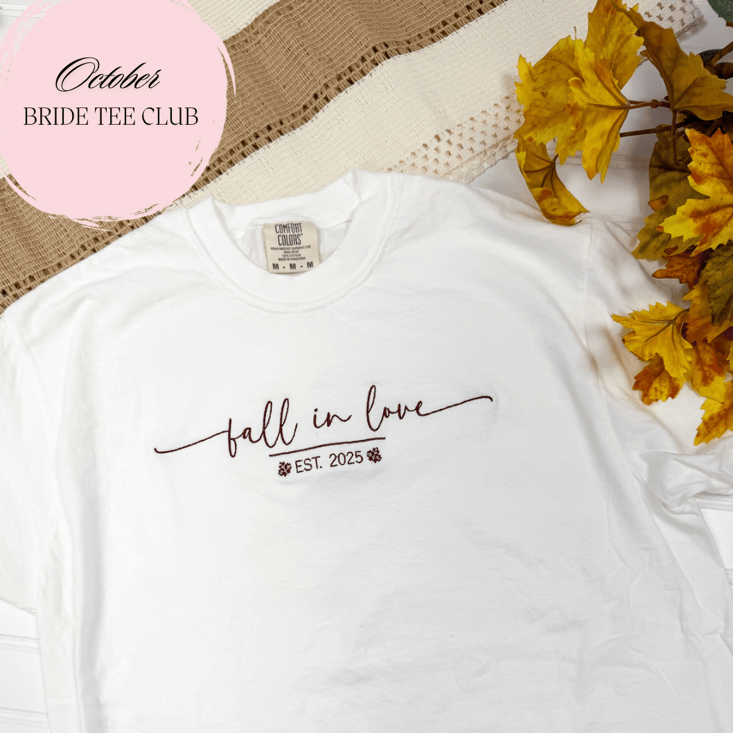 White t-shirt with "fall in love EST 2025" script design and fall leaves on natural fabric background