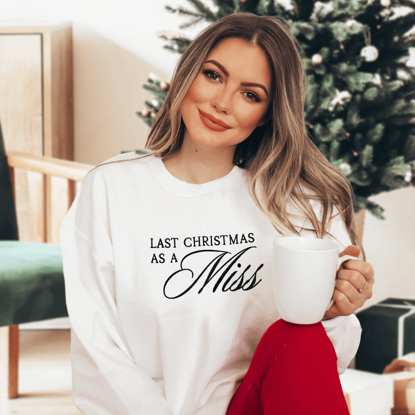 White crewneck sweatshirt with "Last Christmas As A Miss" text in black script font, worn by woman in festive setting with Christmas tree background.