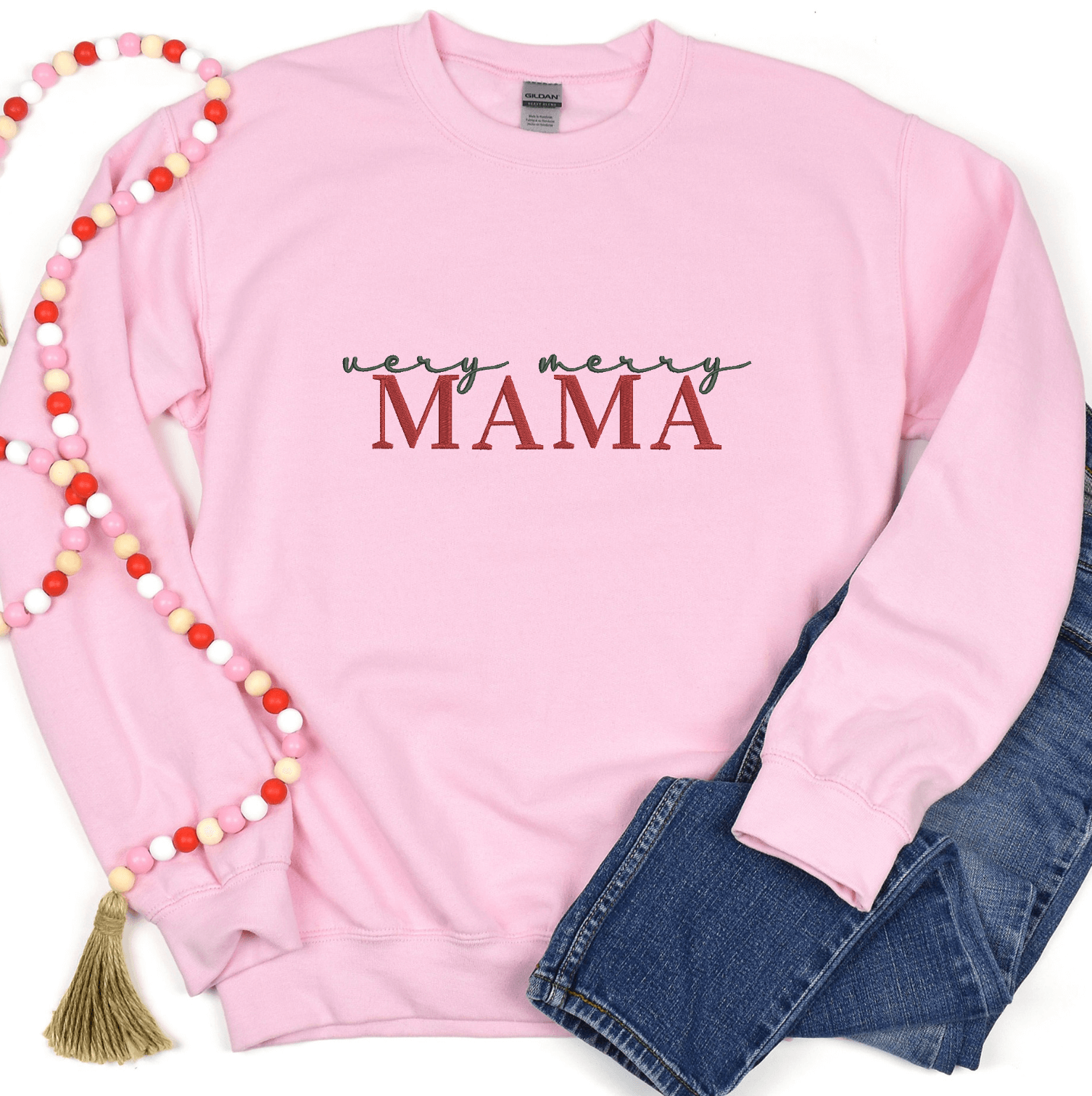Pink crewneck sweatshirt with "Very Merry Mama" embroidered in burgundy text, styled with jeans and a beaded necklace.