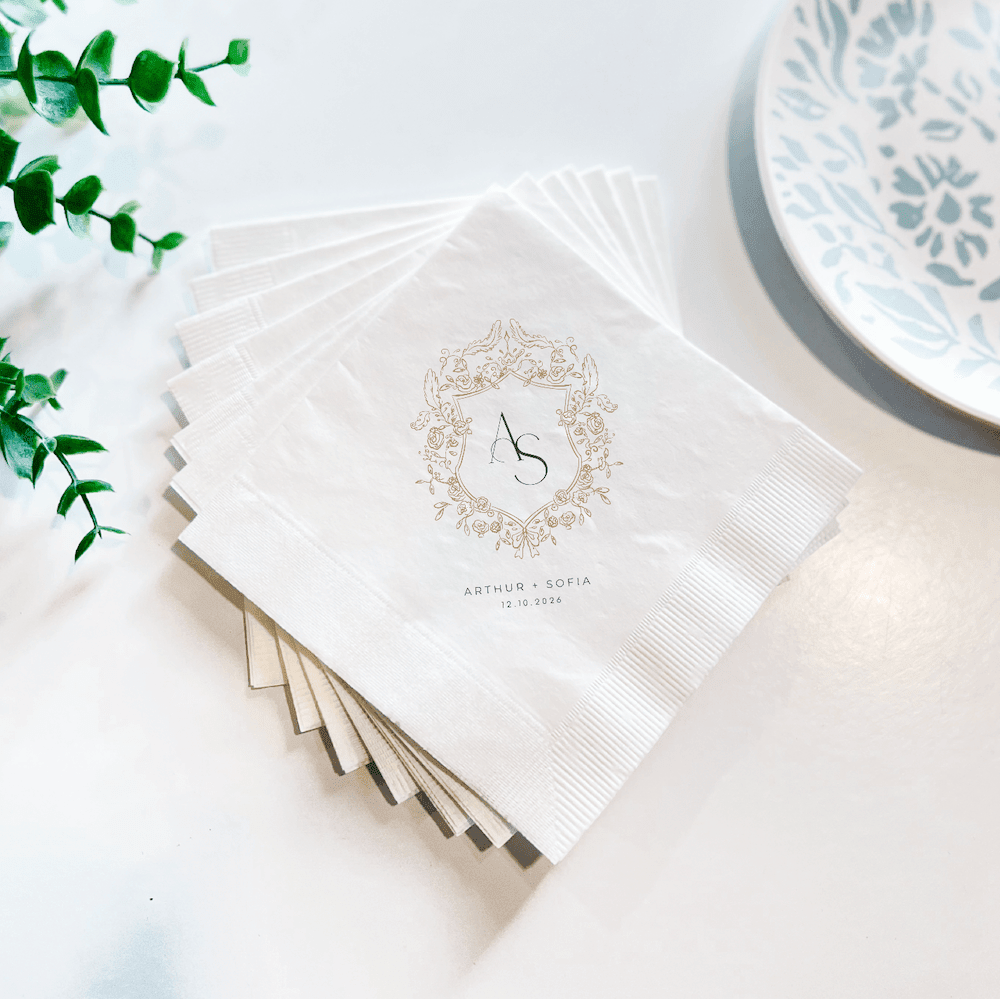 Stack of white napkins with ornate wreath design and personalized names, featuring blue decorative plate and greenery on white surface.