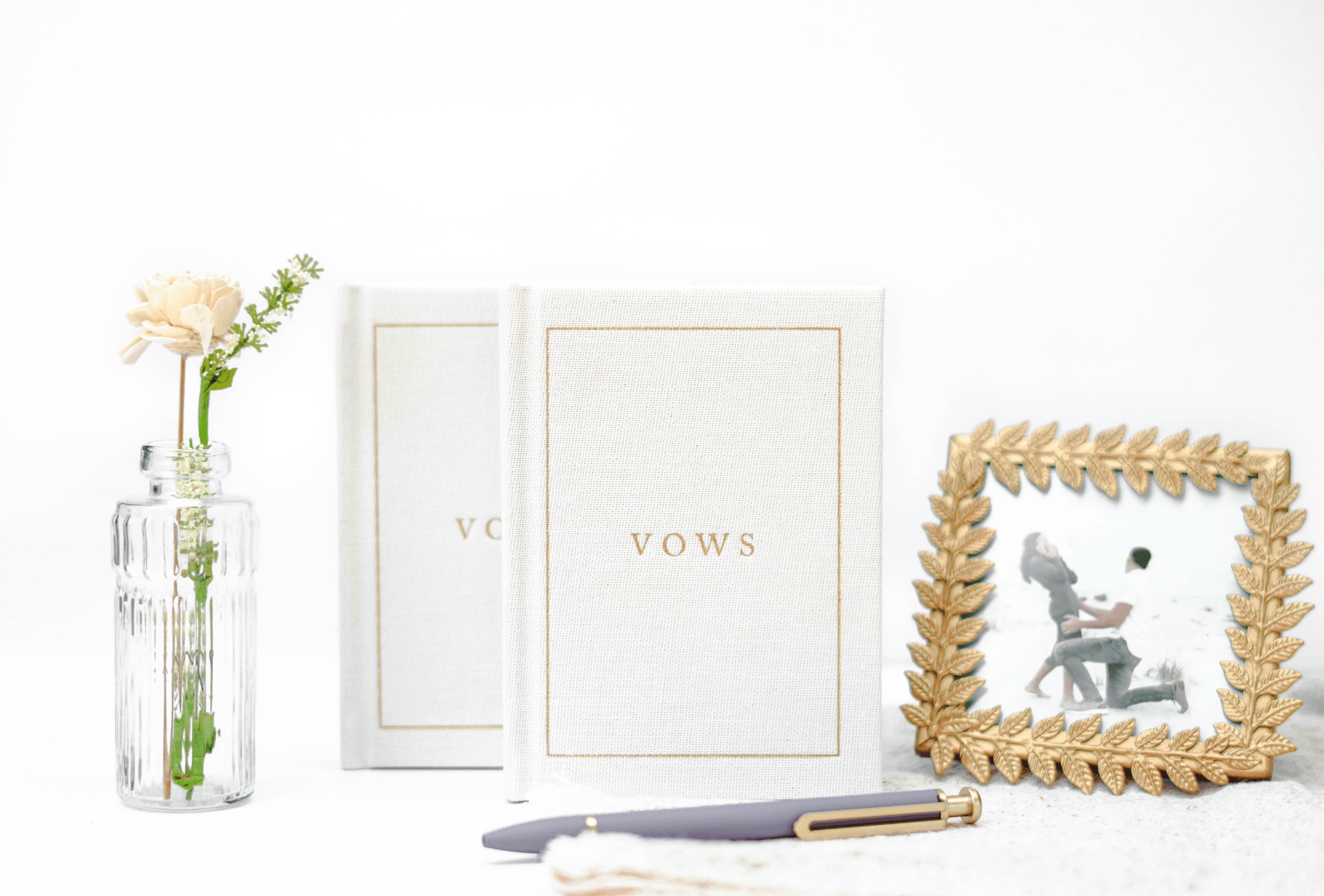 White vow books with gold foil text displayed next to a glass vase with flowers and a gold frame.