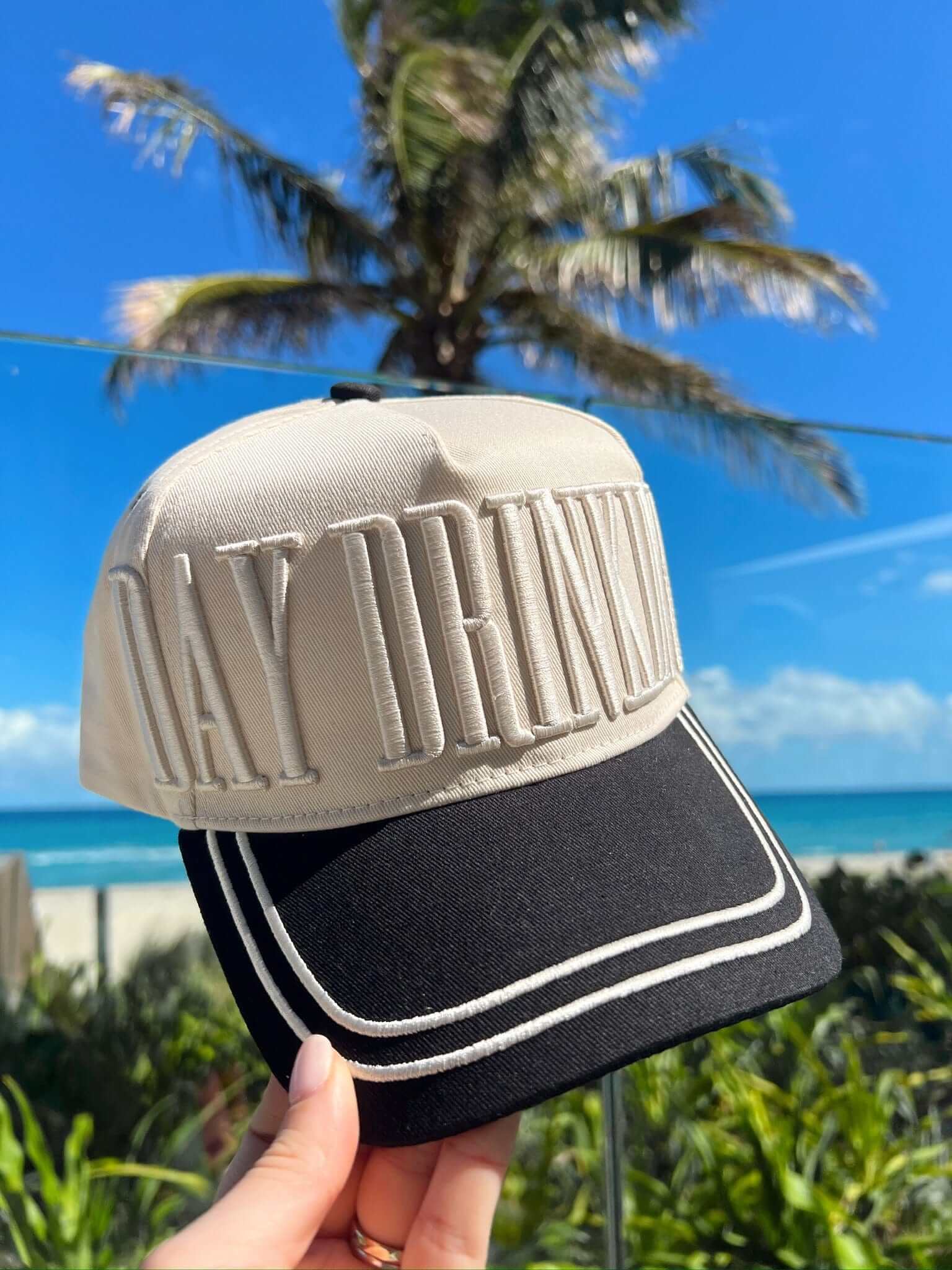 Cream and black vintage trucker hat with "DAY DRINKING" embroidered on the crown, held up against a tropical beach background.